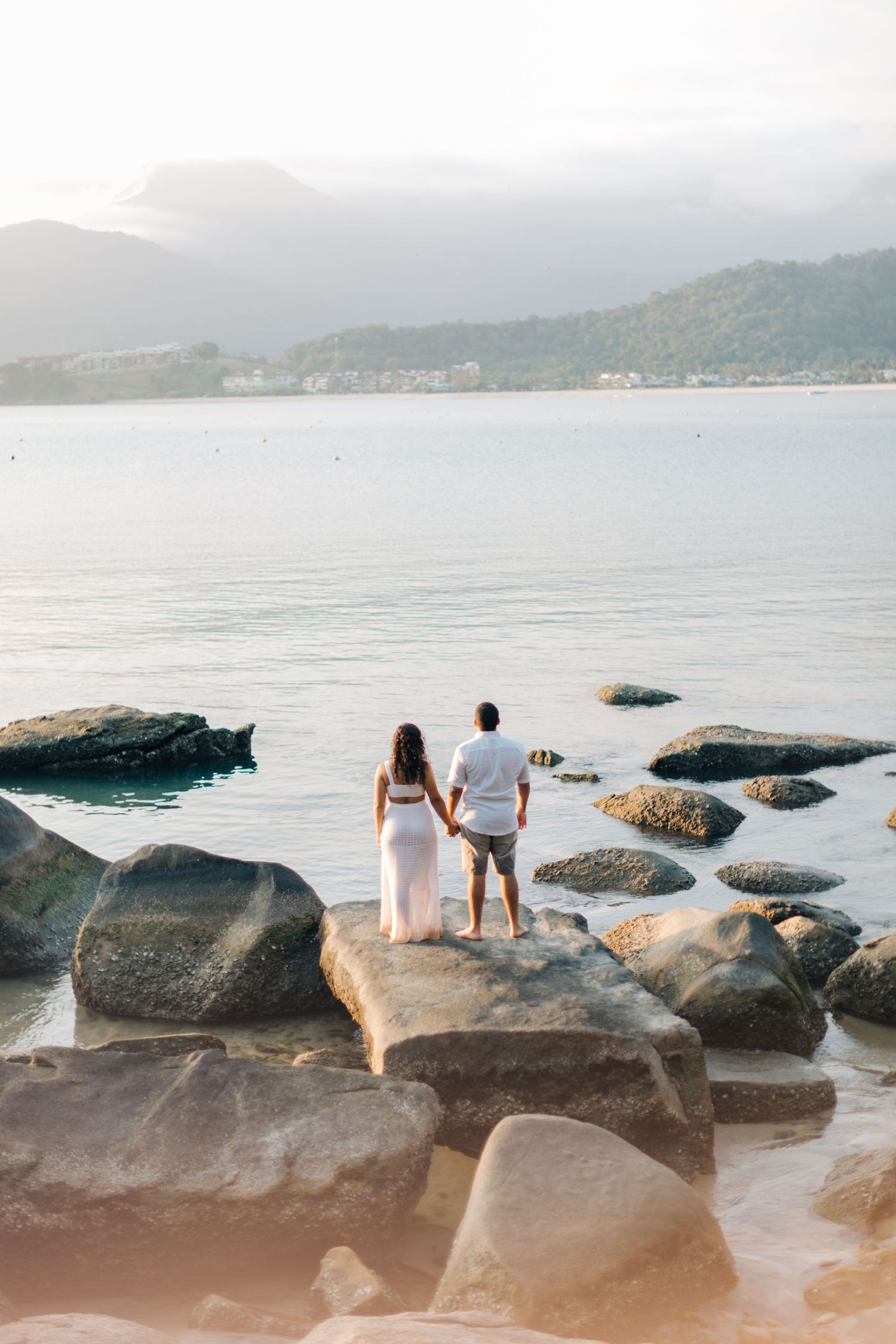 Couple holding hands while standing on rocky beach overlooking misty mountains.