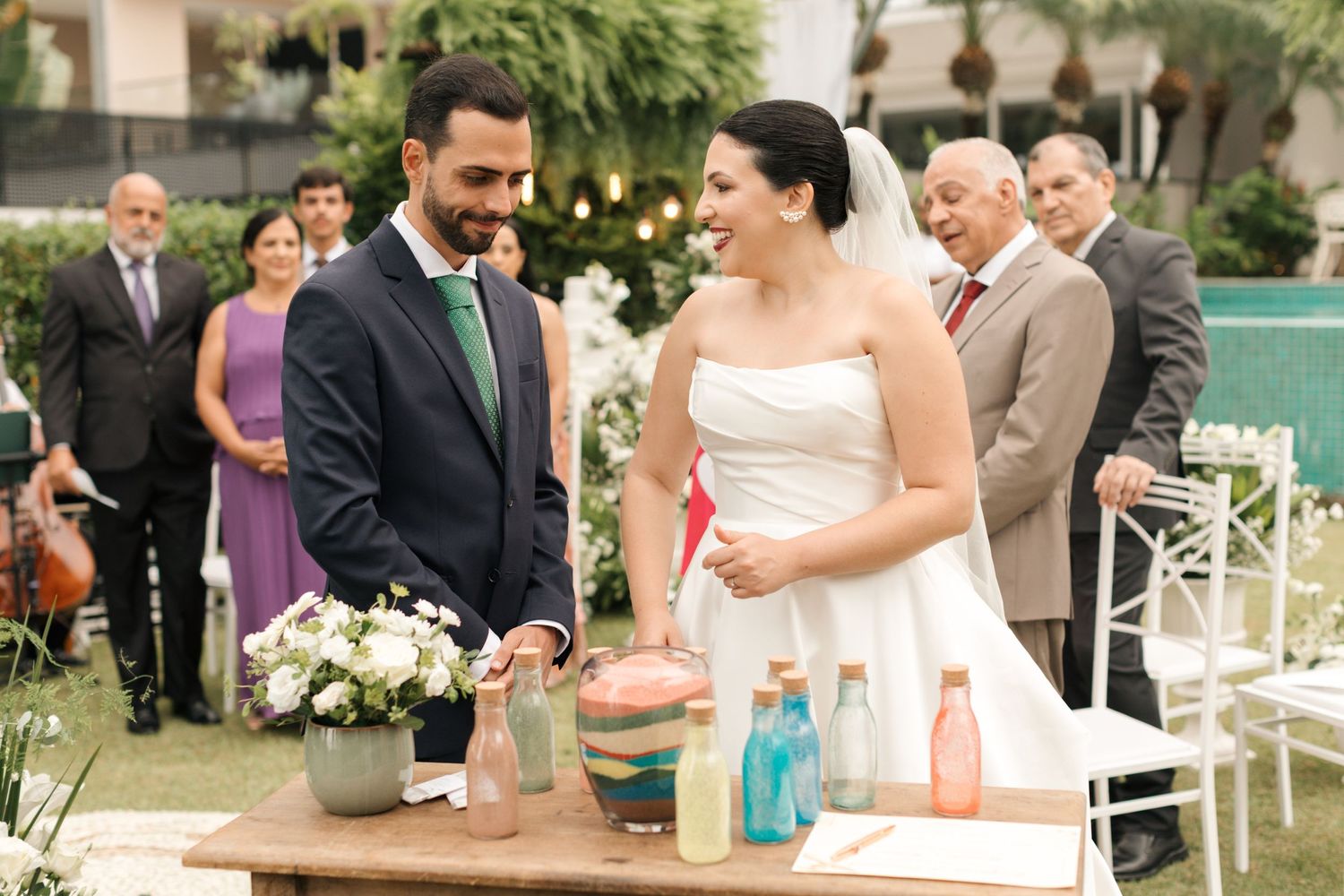 Guests gather around wooden table with colorful beverages at outdoor wedding celebration.
