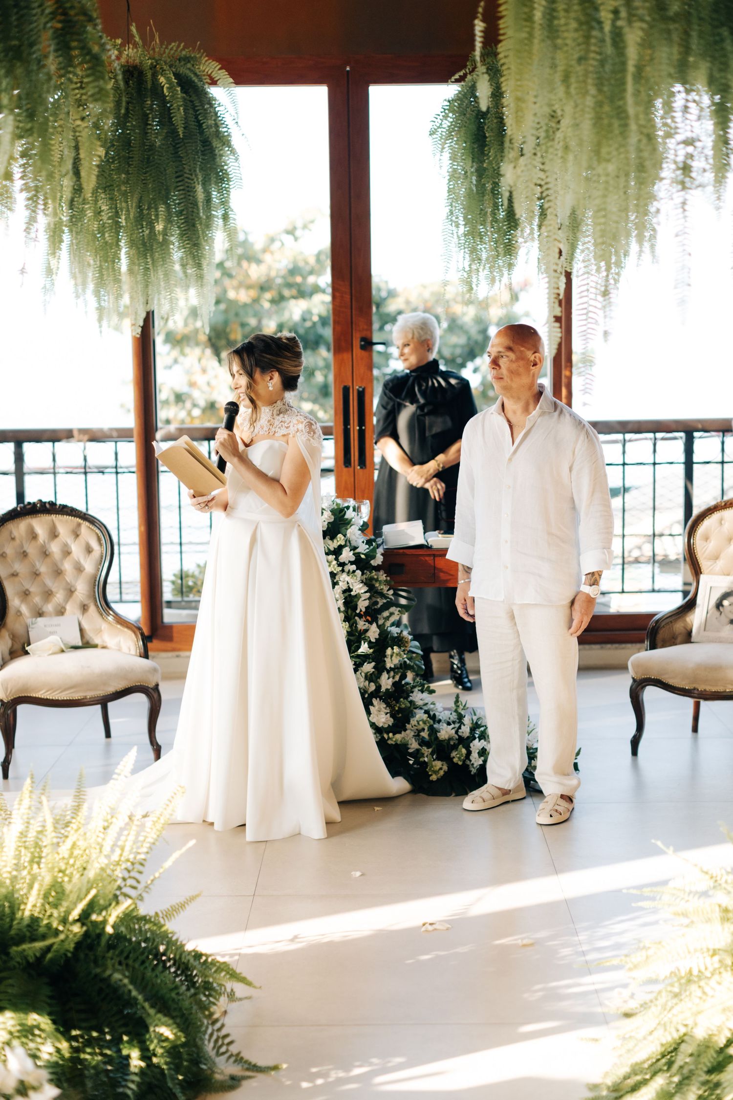 A wedding ceremony in a bright venue decorated with hanging ferns and elegant vintage chairs.