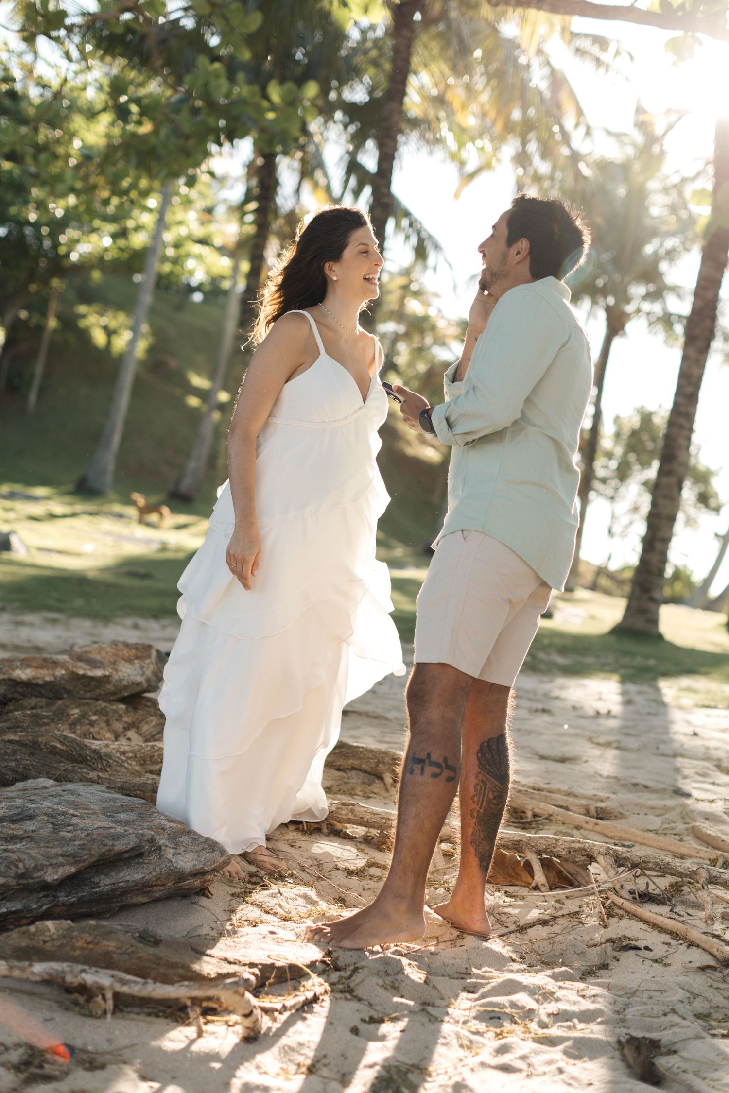 A joyful couple shares a candid moment on rocky terrain during their beach wedding photoshoot at sunset.