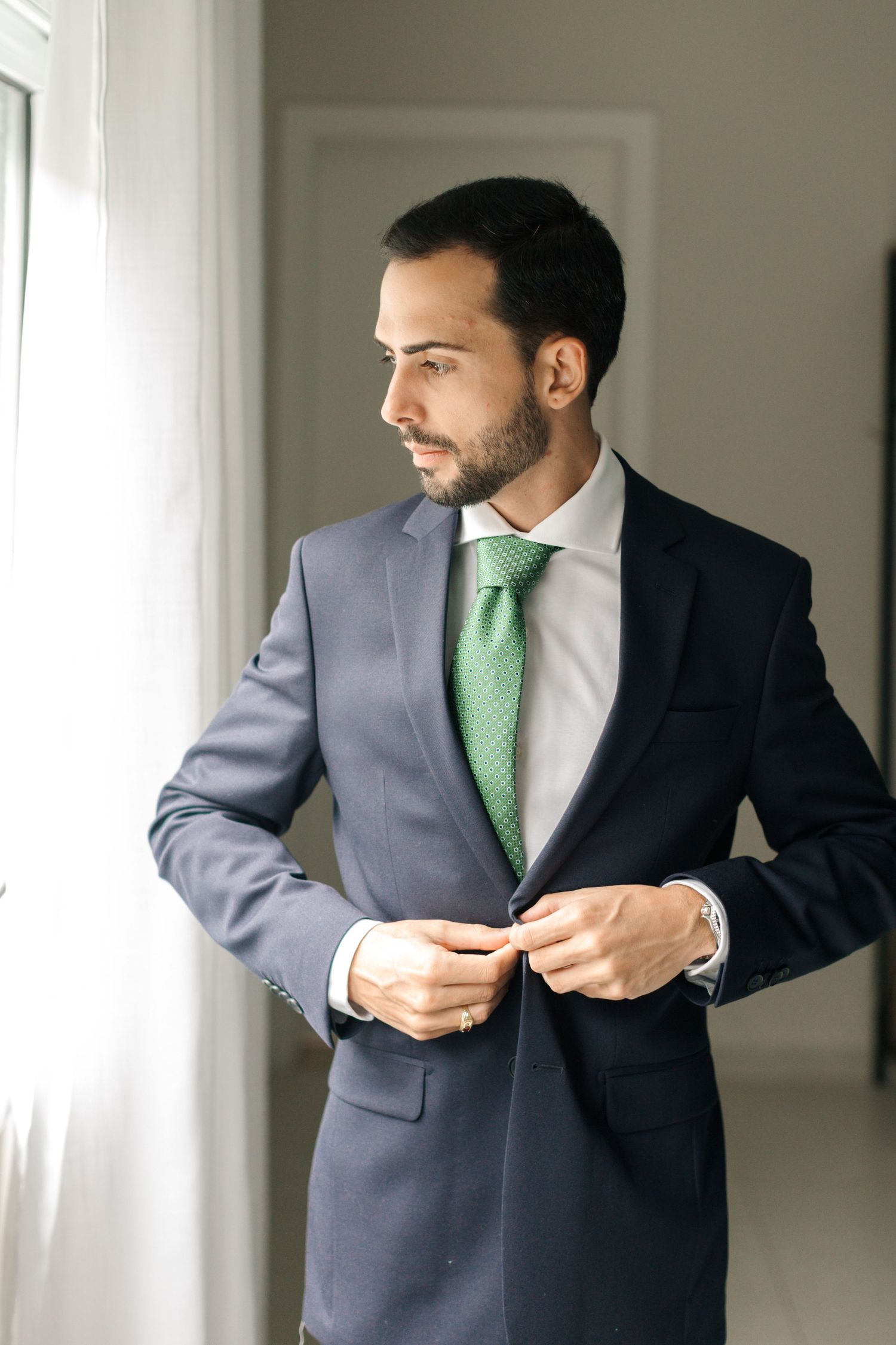 Well-dressed professional in navy suit and green tie stands by window in formal attire.