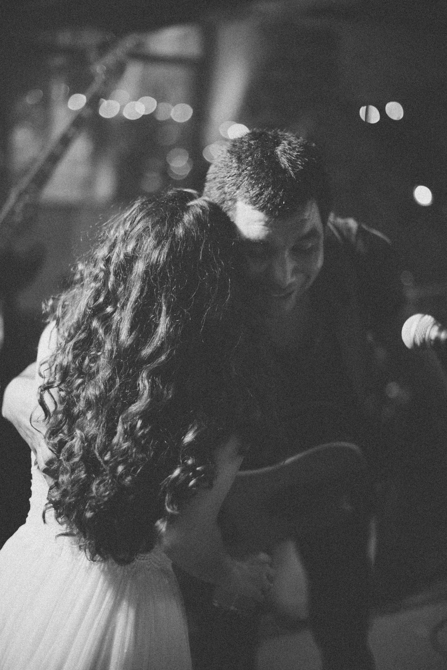 A romantic black and white shot of a couple dancing at an intimate concert venue with bokeh lights in background.