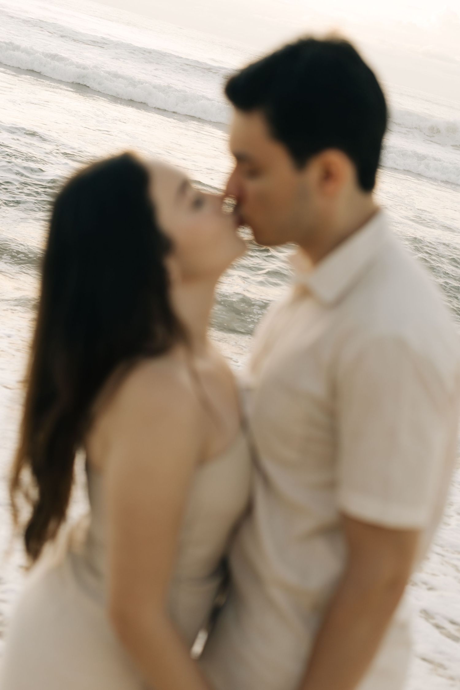 Artistic blurred shot of a couple kissing on the beach with ocean waves in the background.