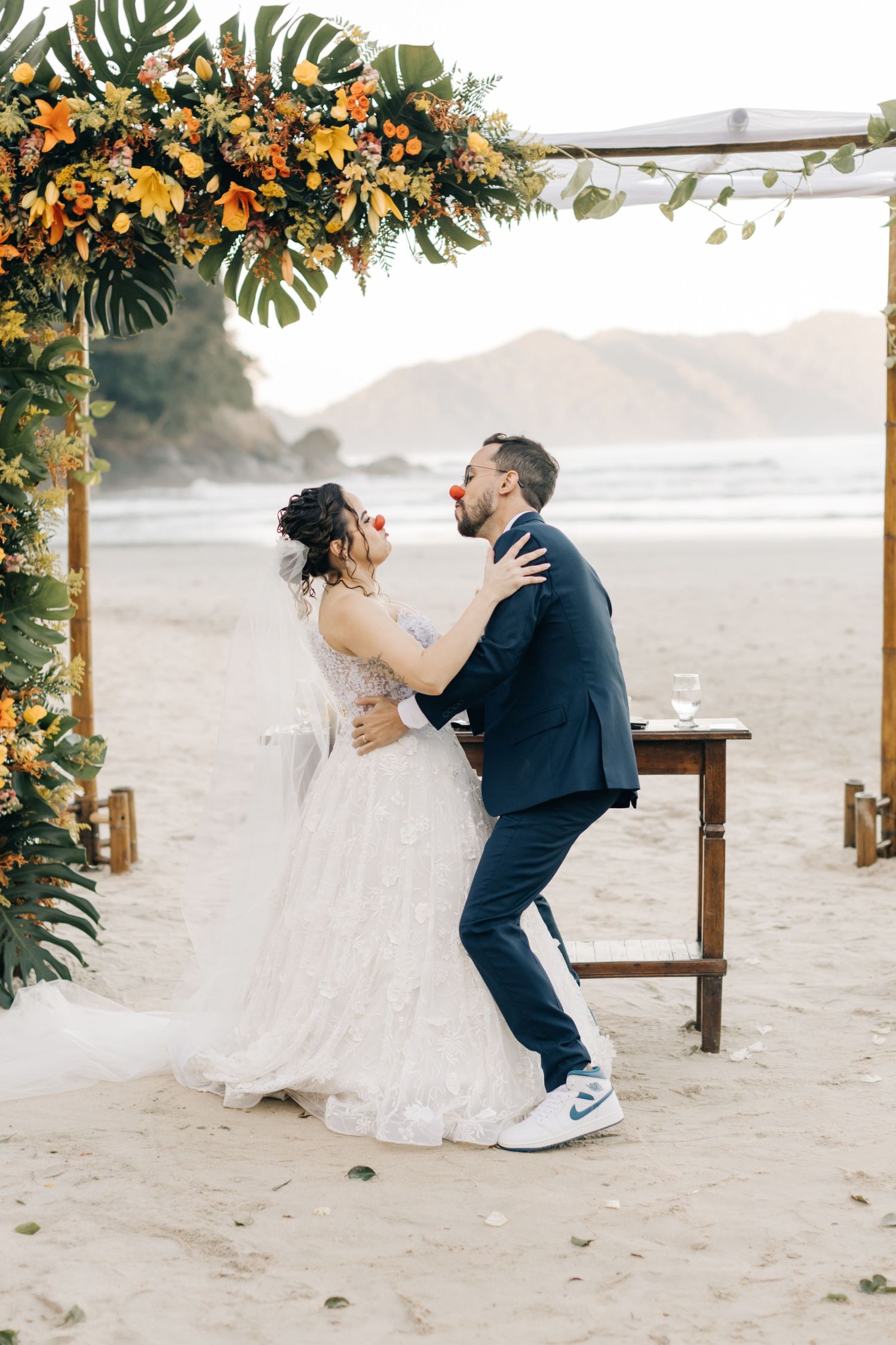 A couple celebrates their beach wedding under a tropical floral arch with orange blooms and monstera leaves.