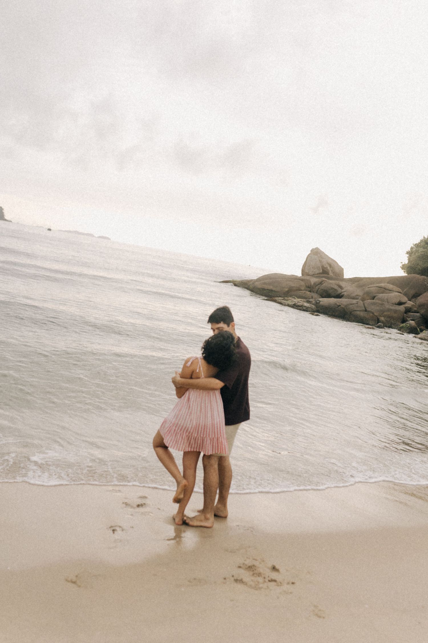 Um casal se abraça em uma praia enevoada com grandes pedras e céu nublado ao fundo.