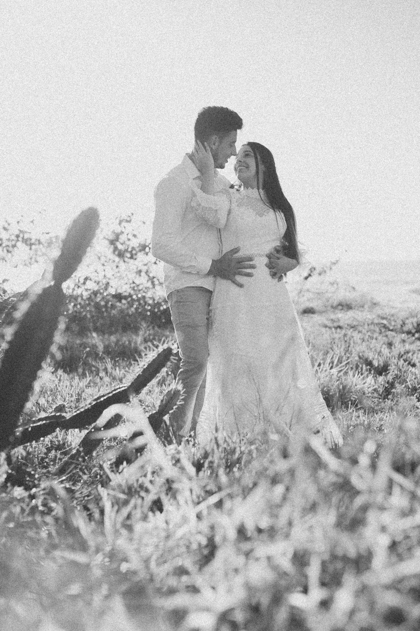 Black and white portrait of couple embracing in desert landscape with cacti silhouettes.