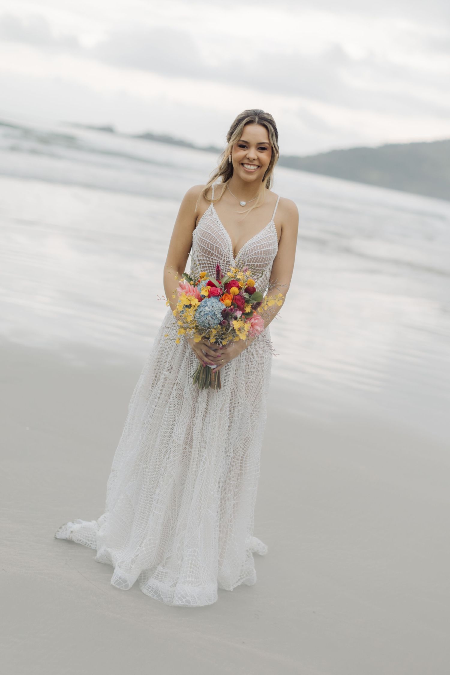 A bride in a white lace wedding dress holds a colorful bouquet while standing on a misty beach.