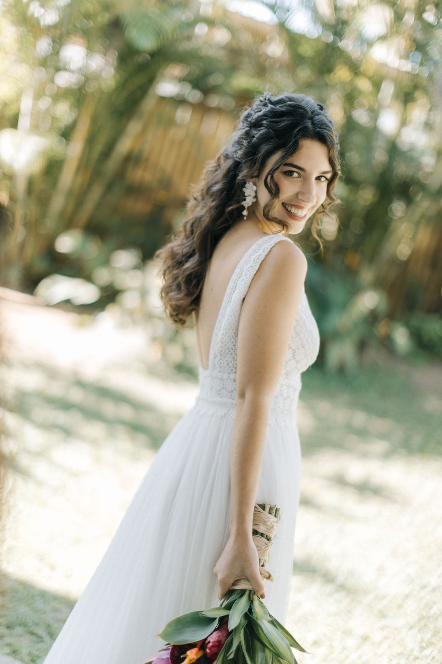 Bride in white lace gown poses gracefully with vibrant tropical flowers outdoors.