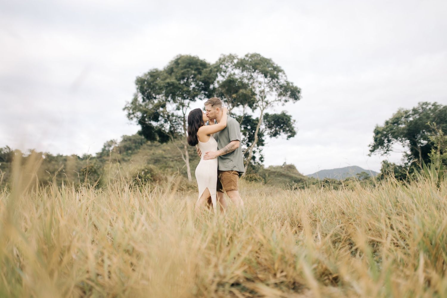 An affectionate couple embraces while standing in tall grass with natural scenery behind them.