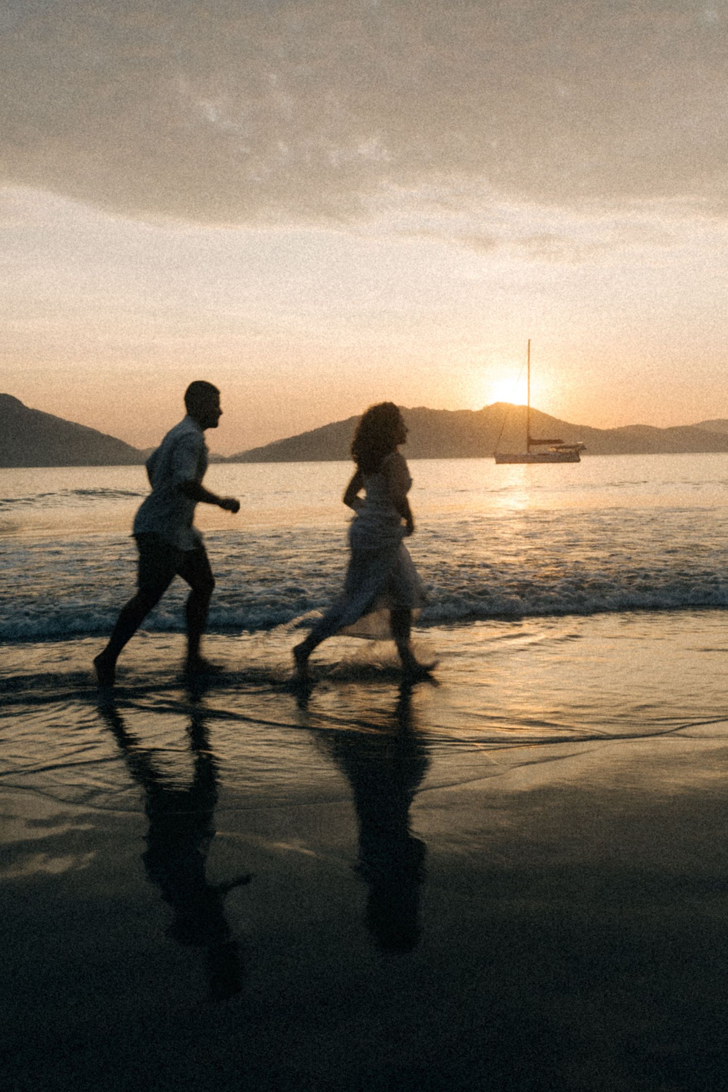 Silhouettes of couple running along beach at sunset with sailboat in distance.