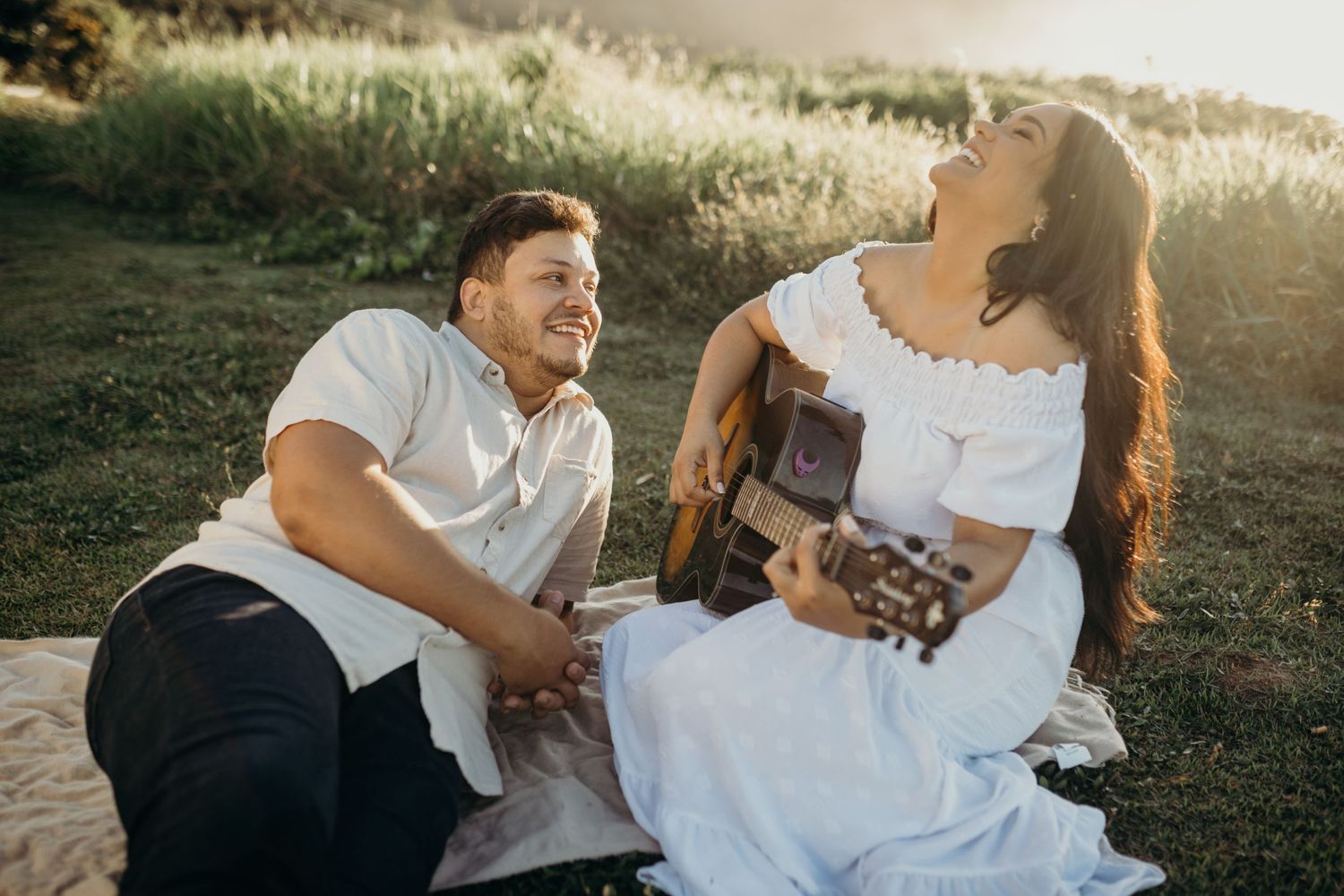 Romantic couple sits on blanket outdoors in sunlight while playing acoustic guitar at sunset.