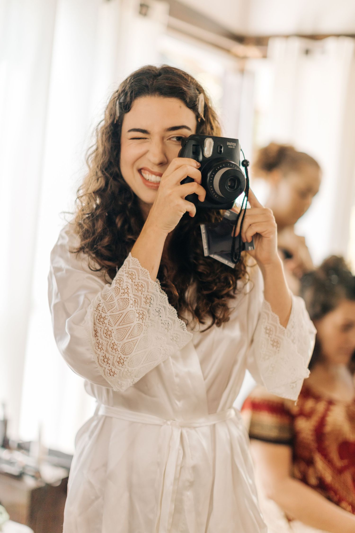 Person in white lace robe joyfully taking photos with instant camera during preparation.