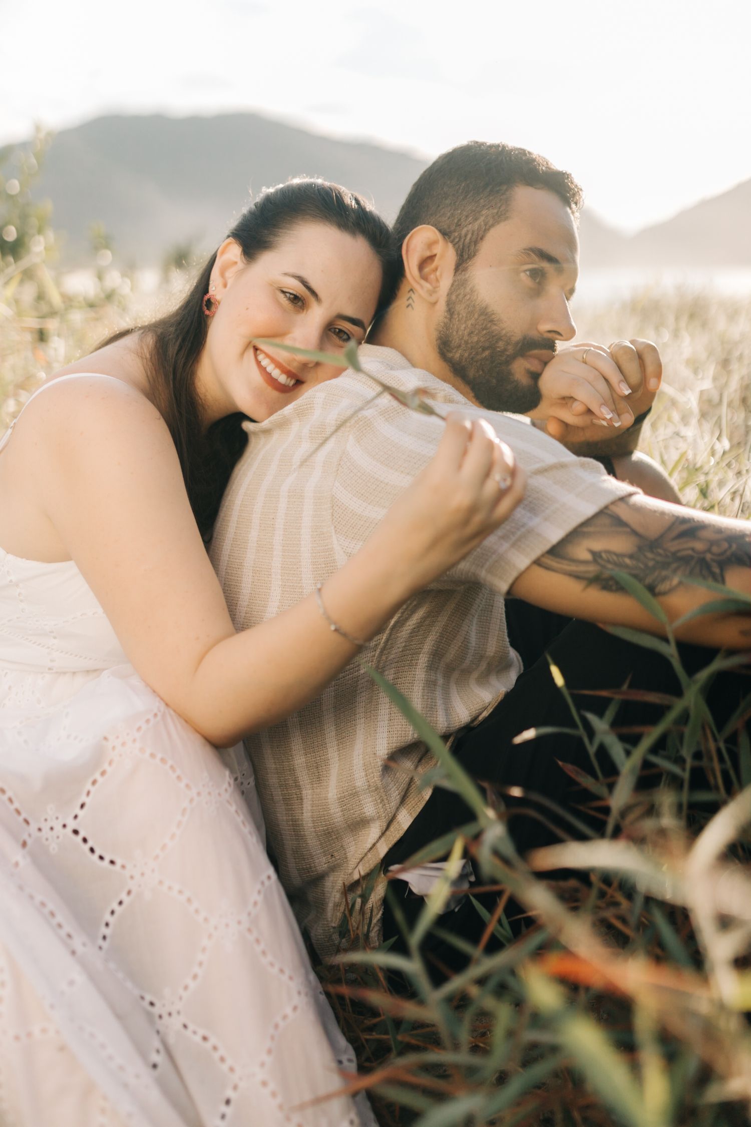 Romantic couple embracing in golden sunlit field with tall grass and mountains in background.