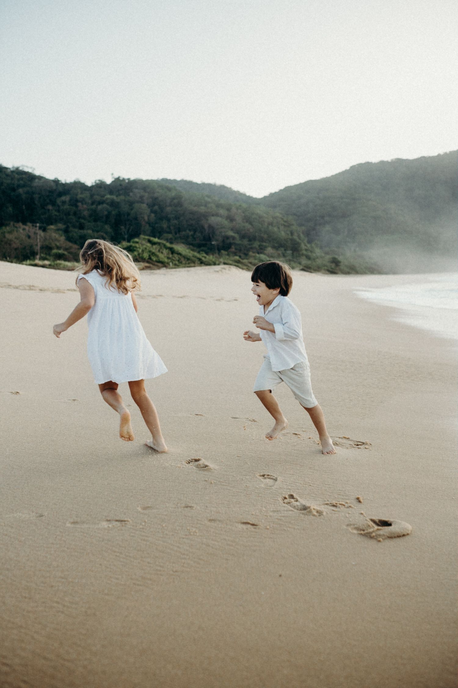 Two children in white outfits running and playing on a misty beach.