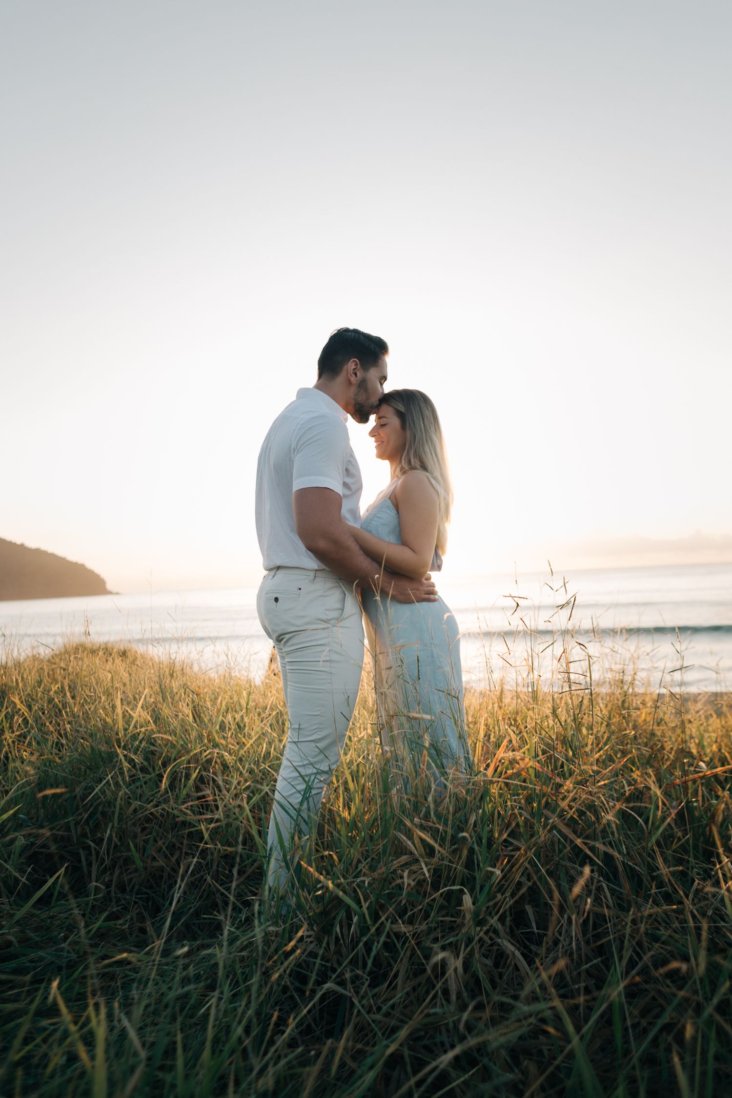 A loving couple stands together in tall grass by the ocean during a golden sunset.