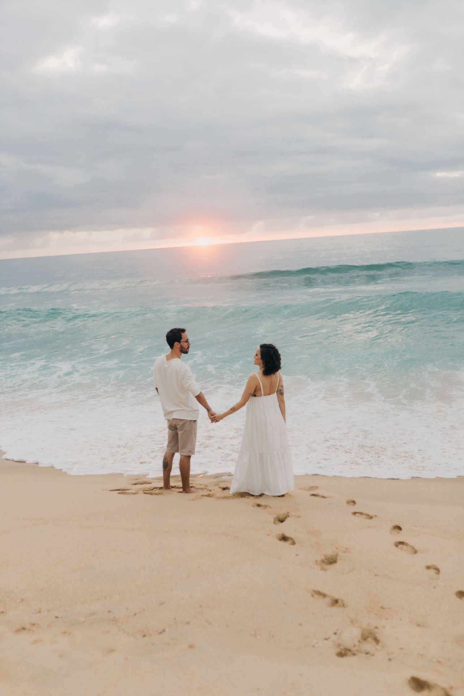 A couple in white clothing walks hand in hand along the shoreline during golden hour.