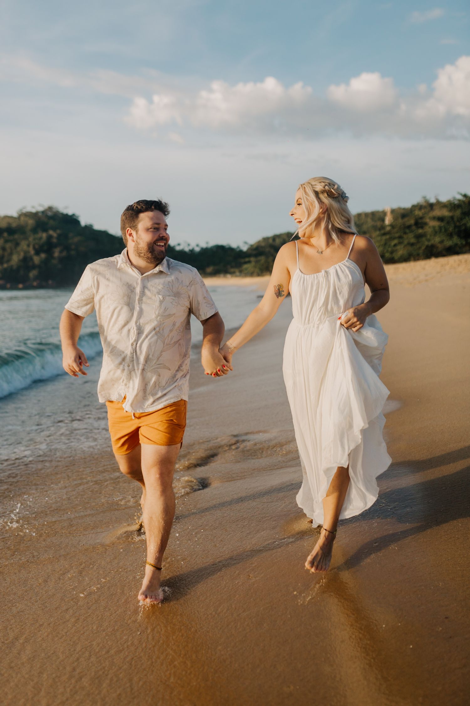 Joyful couple running barefoot along ocean shoreline in coordinated white and orange outfits.