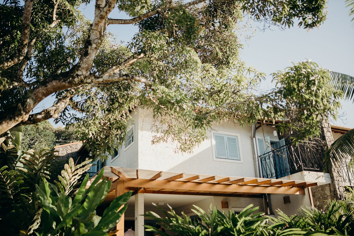 White stucco building with wooden pergola surrounded by lush tropical landscaping and trees.
