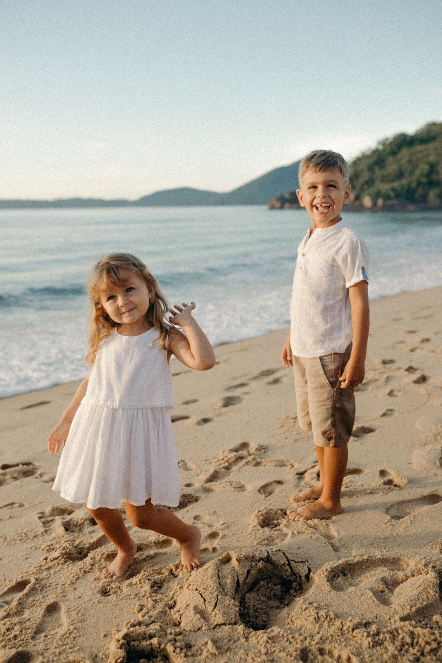 Two children playing on a scenic beach at sunset with mountains in the background.