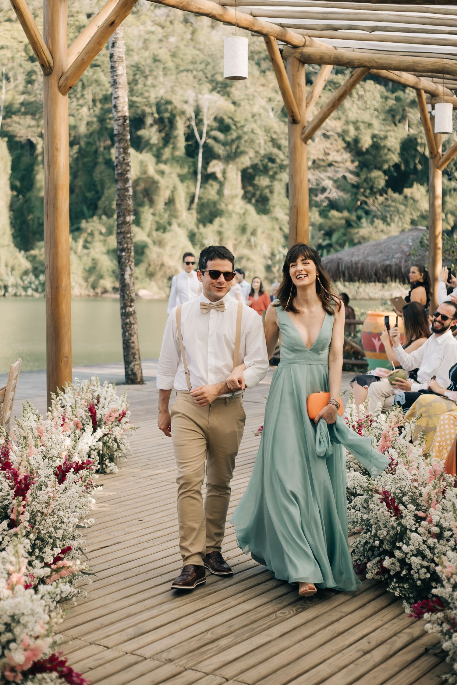 Formal event guests walking along flower-decorated wooden pathway by lake.