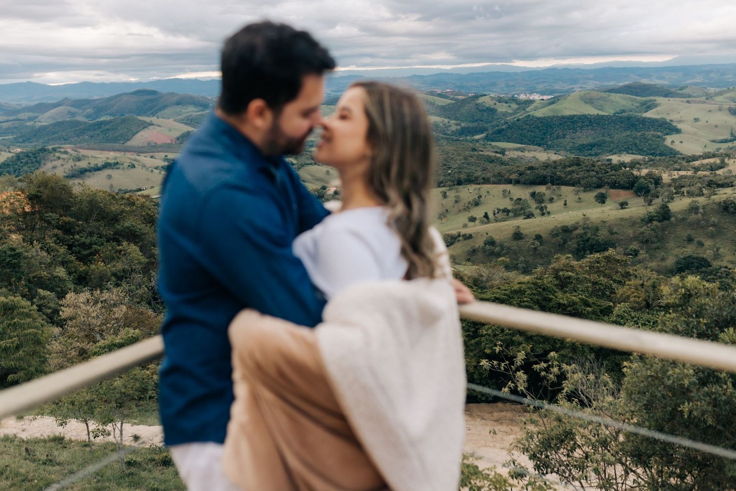 A couple embraces on a mountaintop deck with panoramic views of green rolling hills.