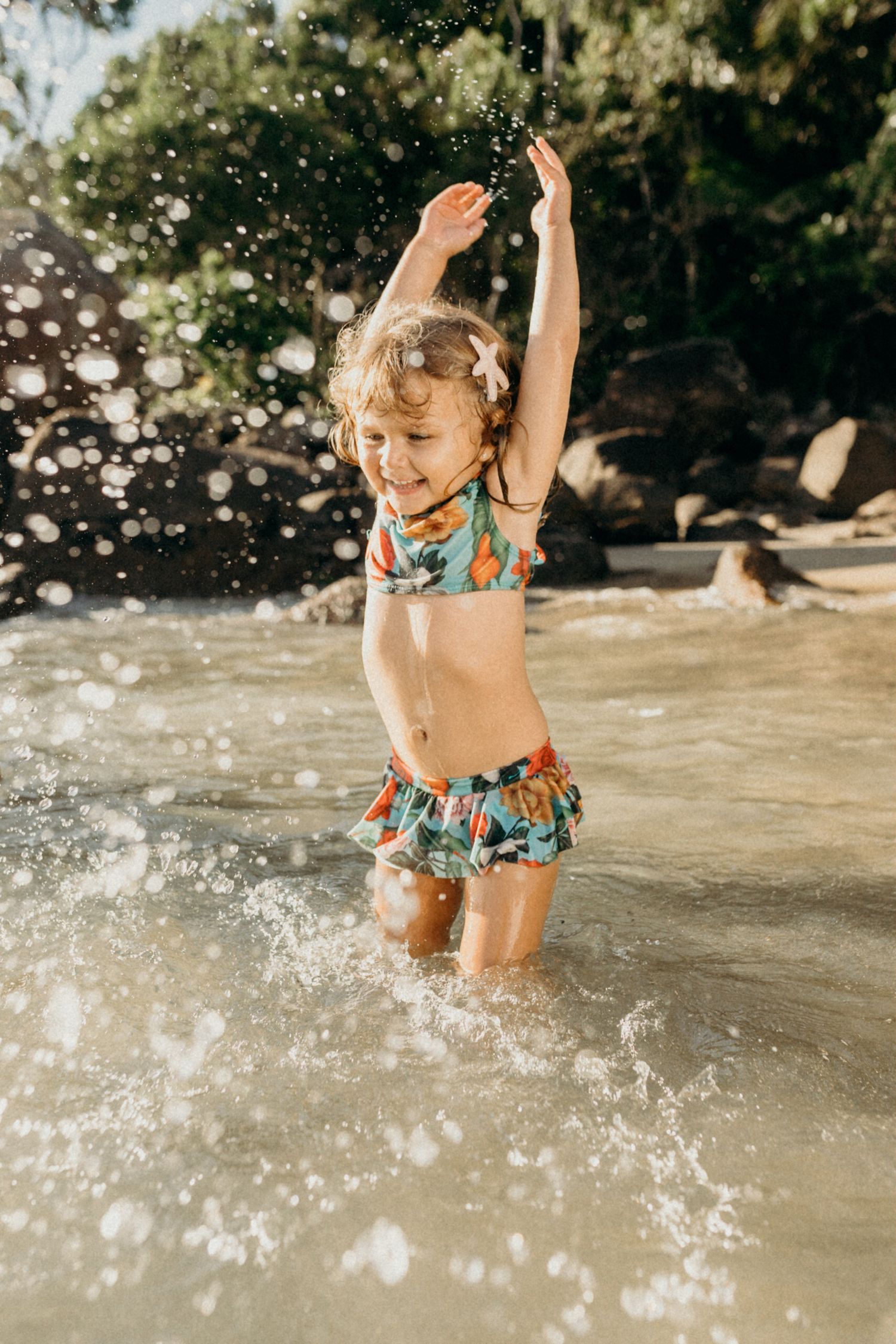 Young child in floral swimsuit jumping and splashing in ocean water with sunlit sparkles.