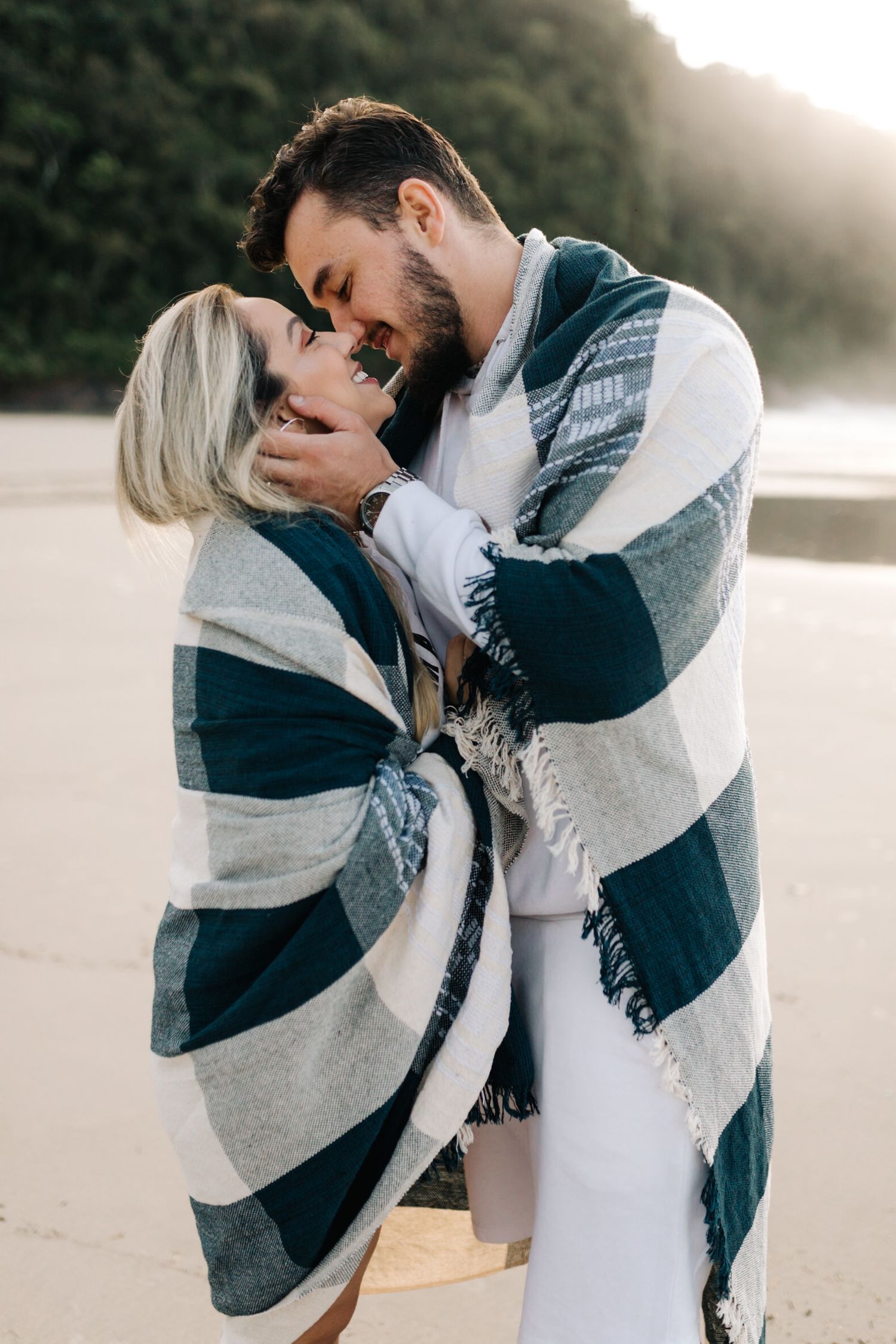 A couple smiles while cuddling under a navy and white striped blanket on the beach.