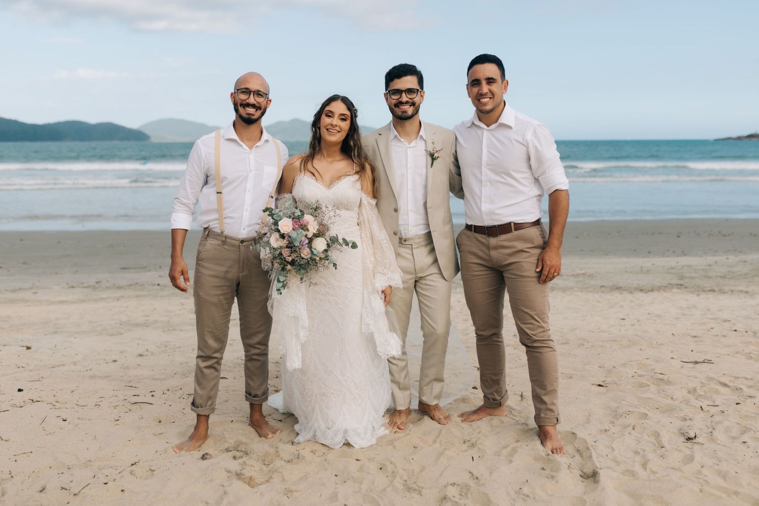 Wedding party poses for photos on beach with mountains and ocean in the distance.