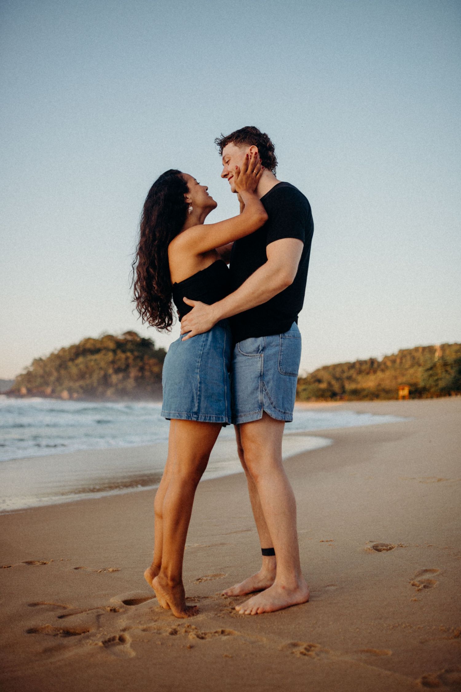 Couple embraces lovingly on beach at sunset with coastal cliffs in background.
