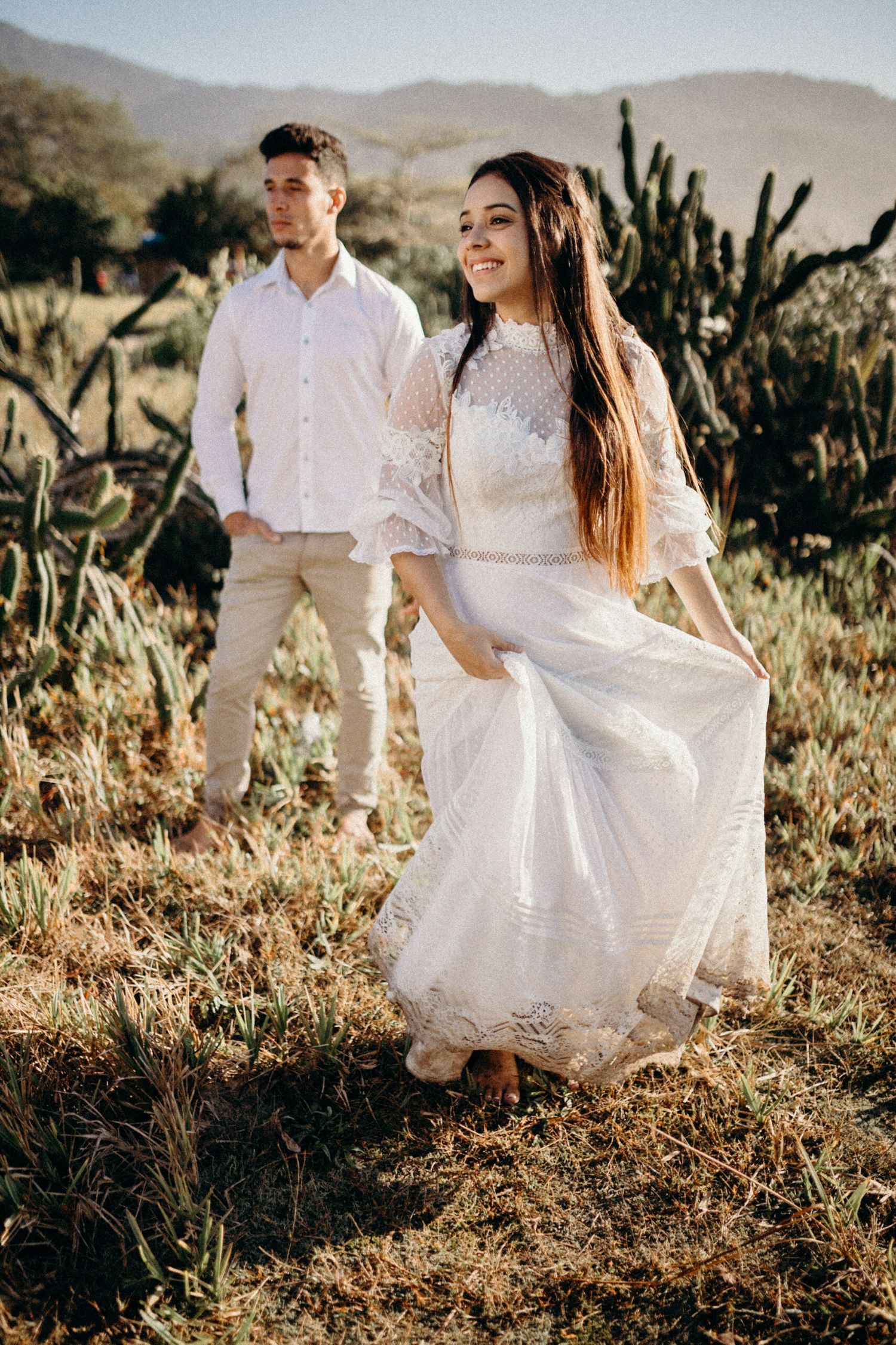 A couple in white outfits poses in a desert landscape with cacti and mountains in the background.