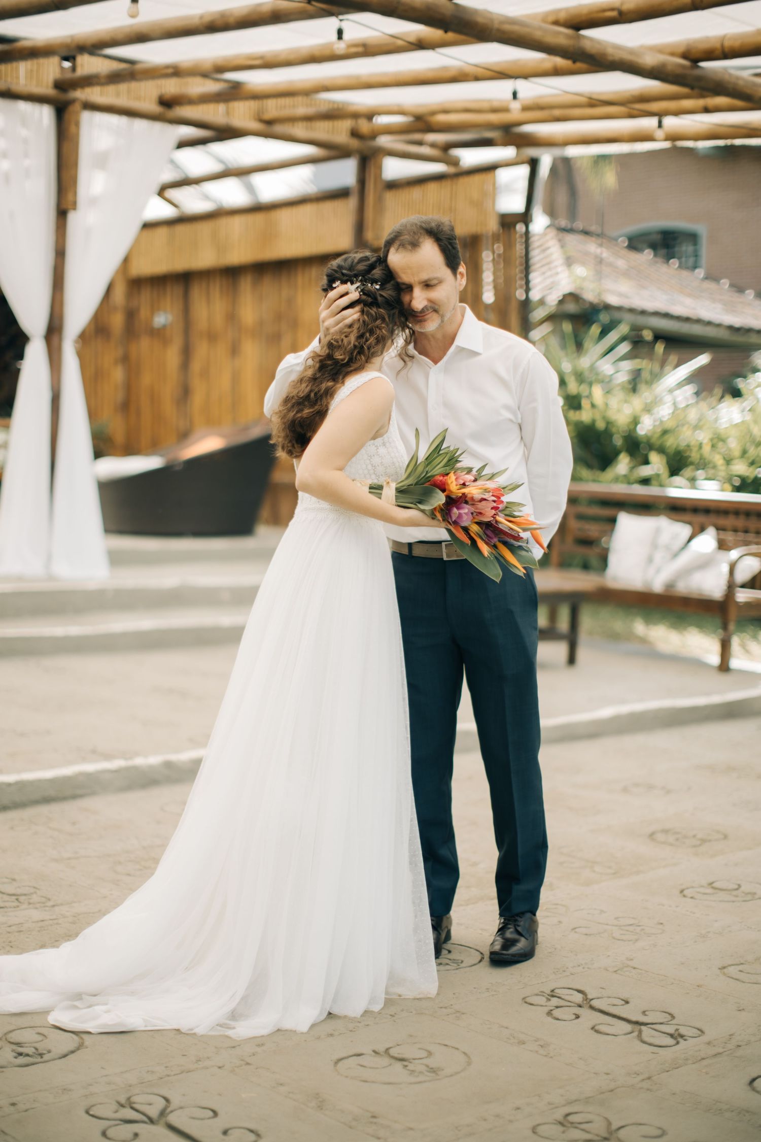 A newlywed couple embraces under a wooden pergola during their tropical wedding celebration.