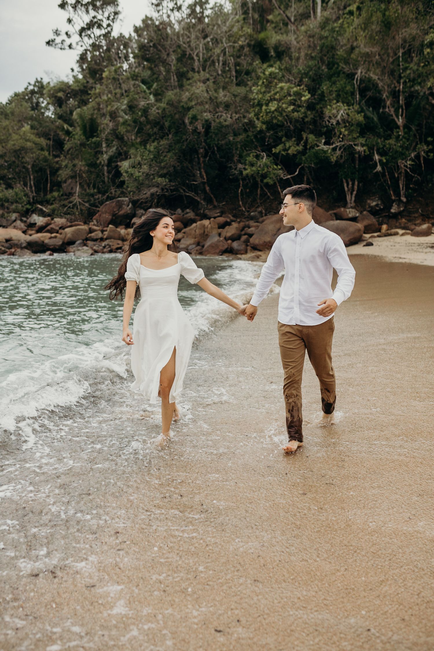 A couple runs playfully through incoming waves on a scenic beach surrounded by trees.