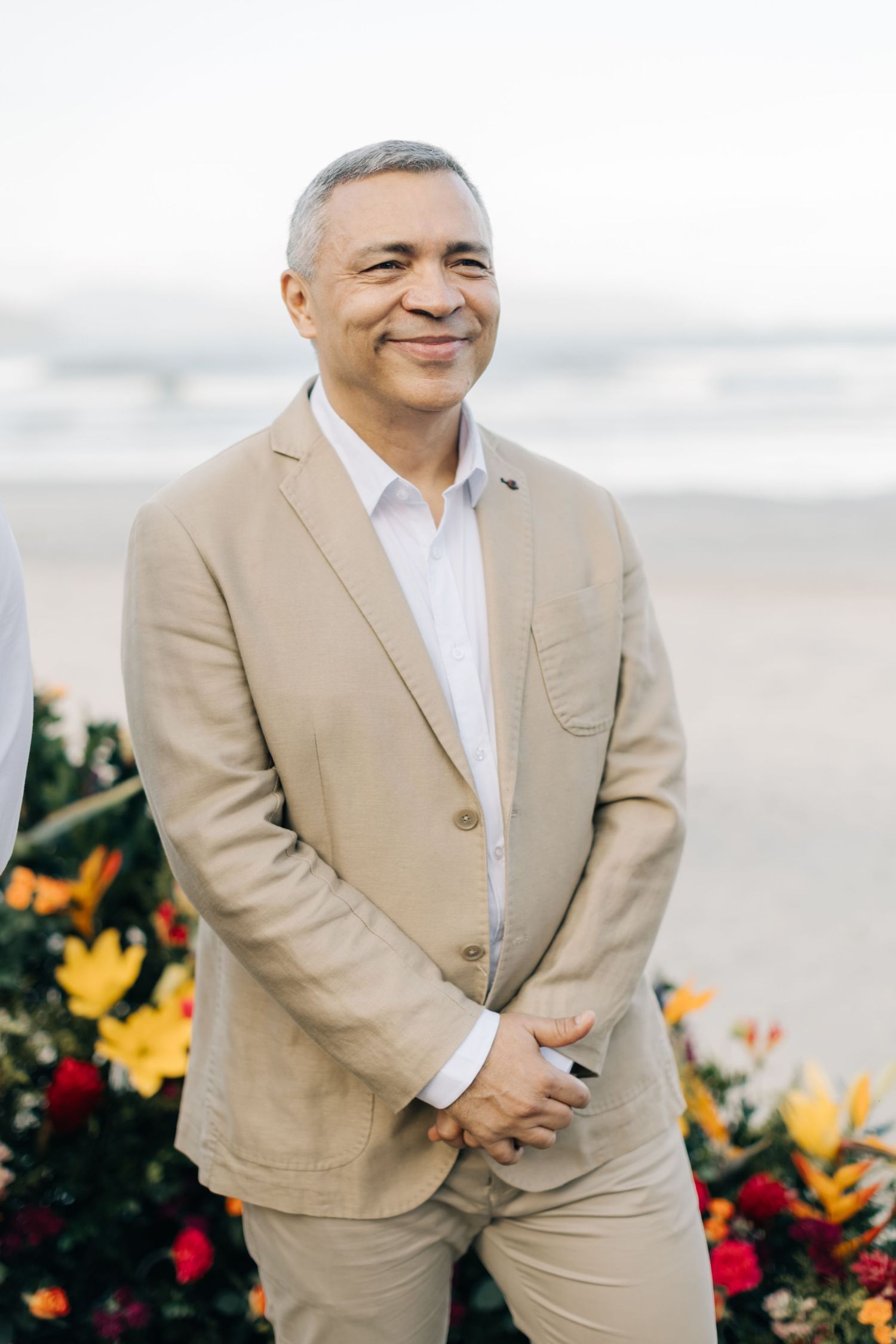 Portrait of person in tan suit smiling against beach background with floral decorations.