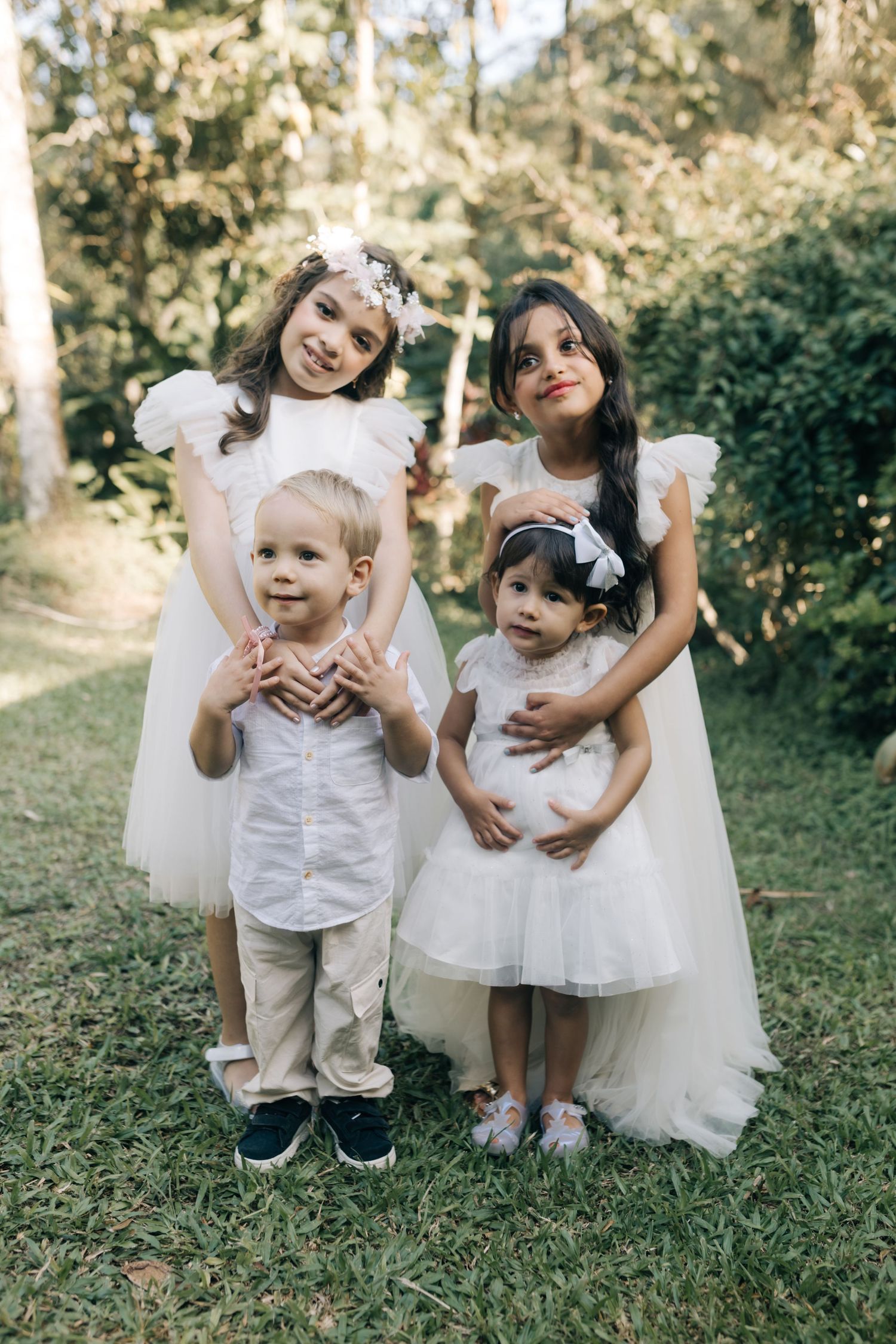 Young children in white outfits play and pose together on grass at a wedding.