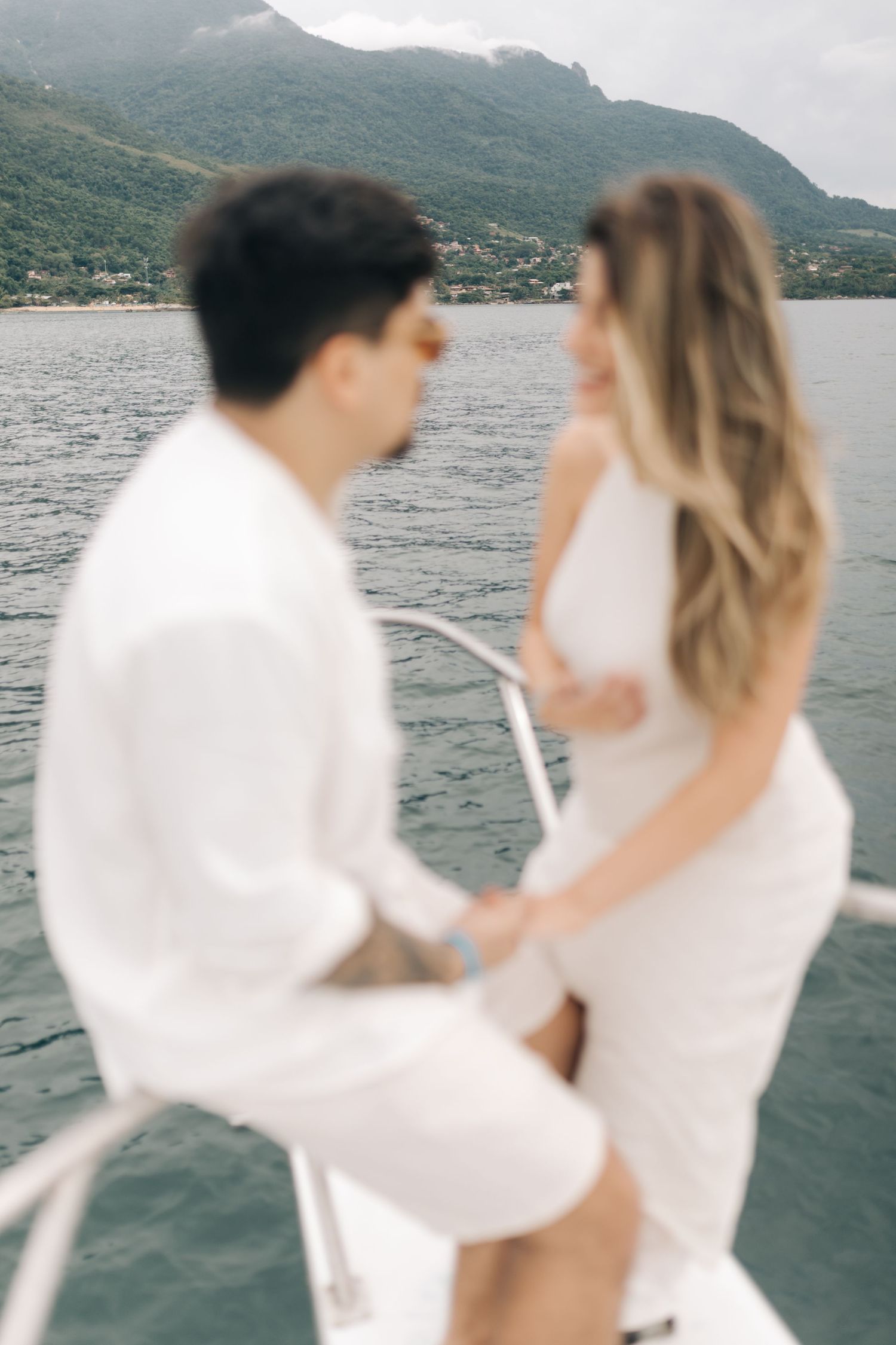 Couple in white outfits sharing intimate moment on boat with mountains in background.