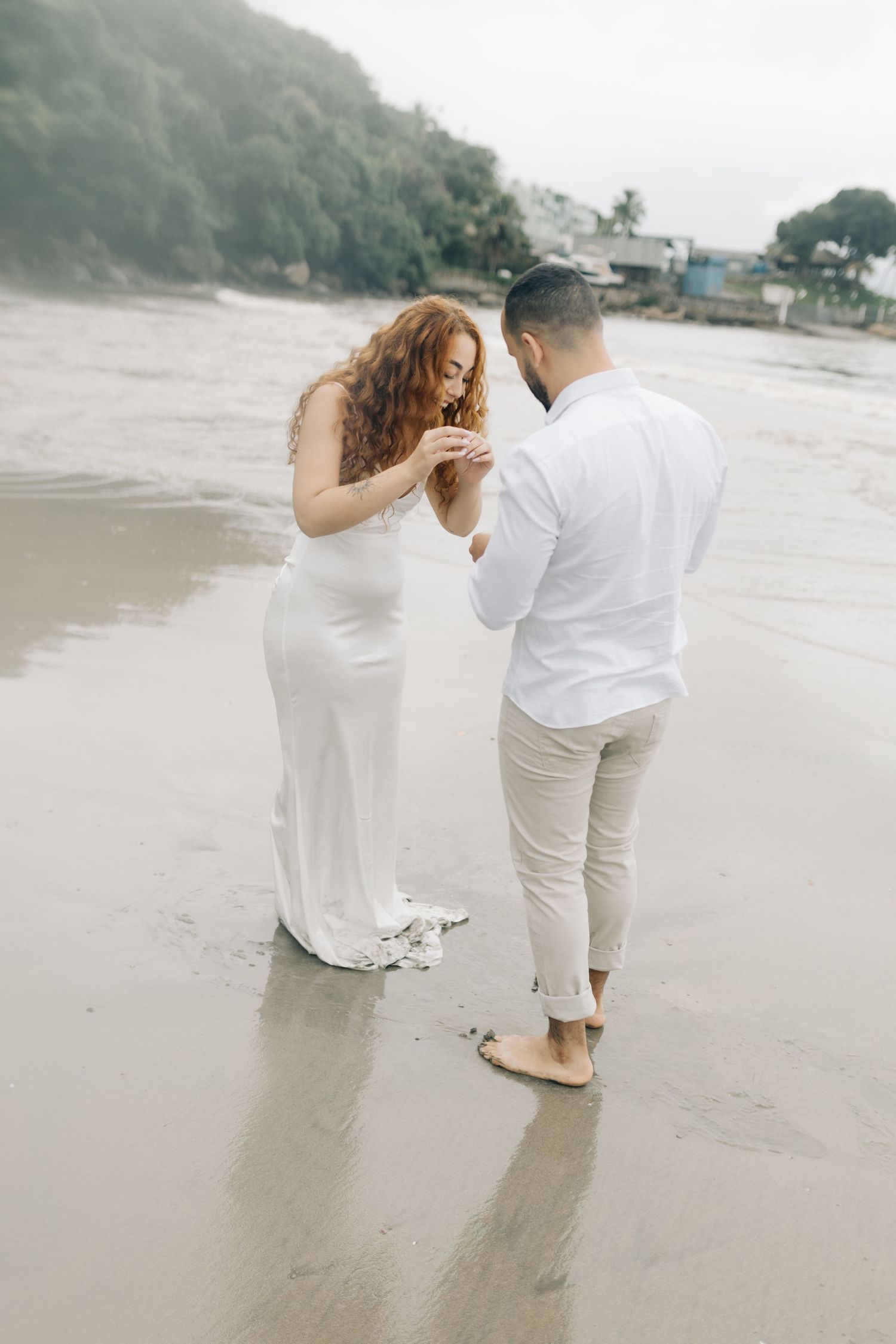Two people in white clothing playfully interact while strolling on a sandy beach.