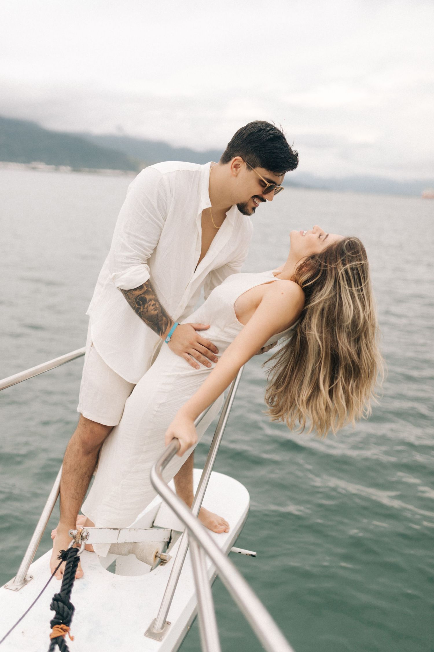 A romantic couple in white outfits share an intimate moment on a boat with mountains in the background.