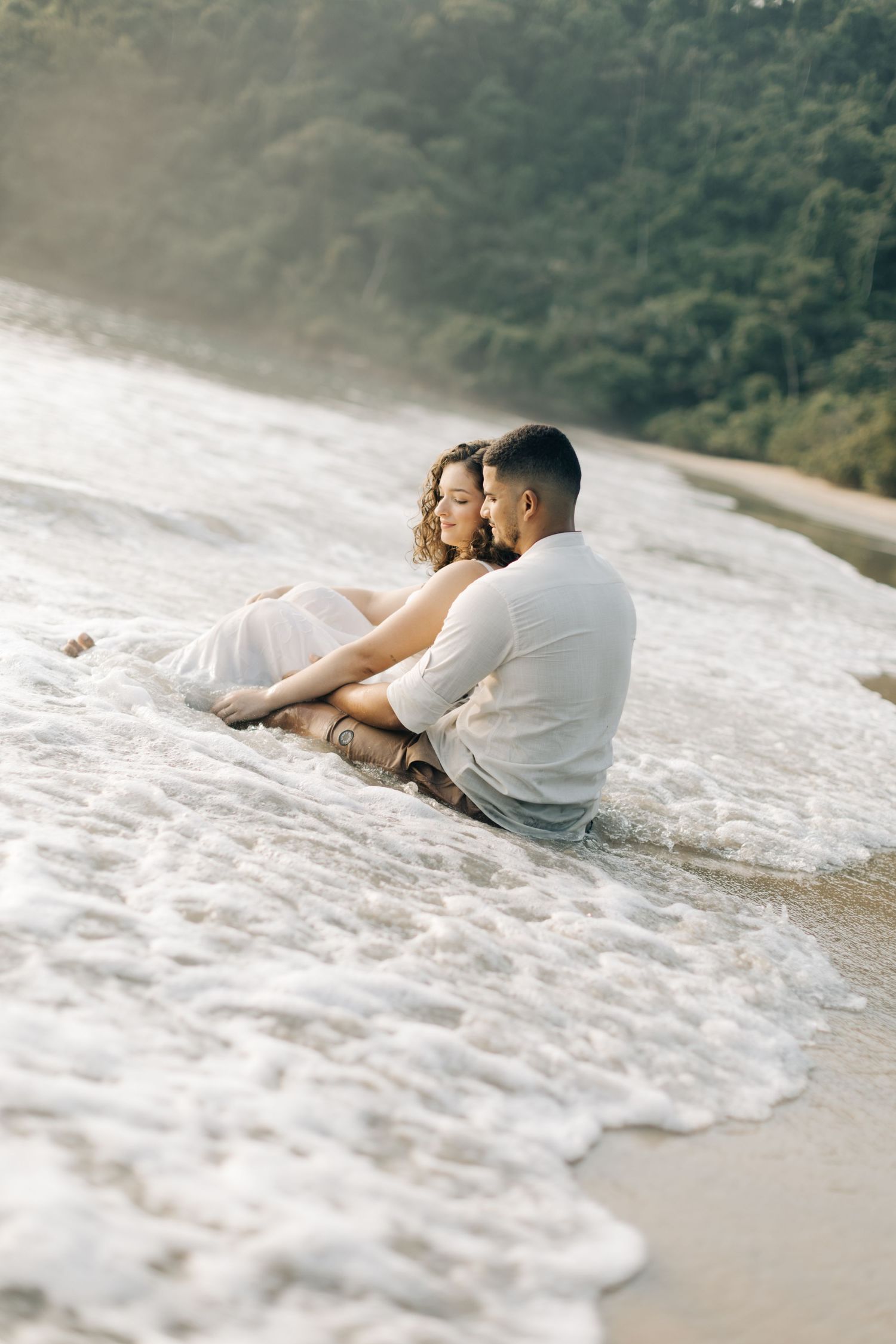 A couple sitting together on a beach as waves wash over their feet.