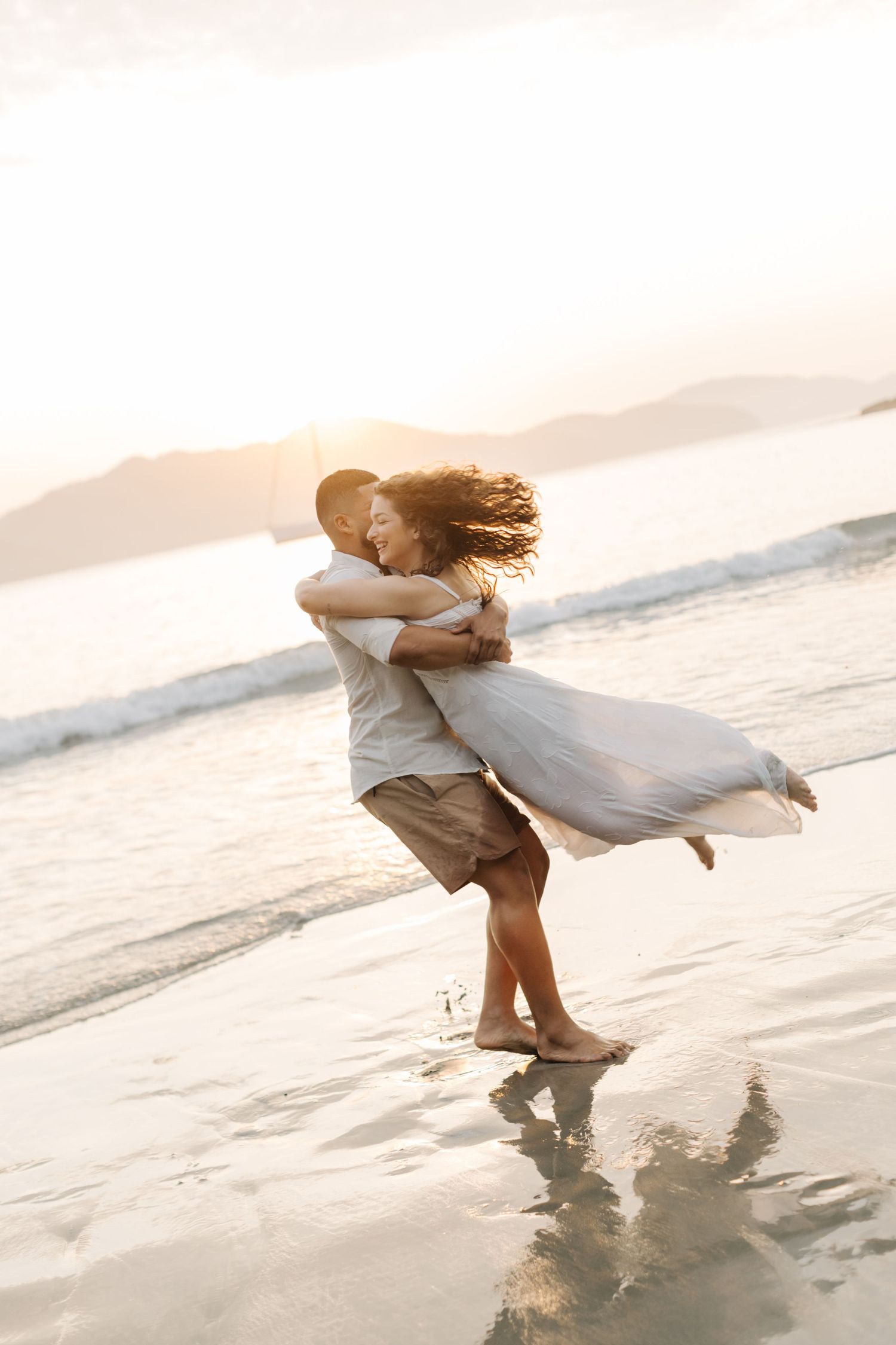 Romantic couple spinning on beach with flowing dress during sunset photoshoot.