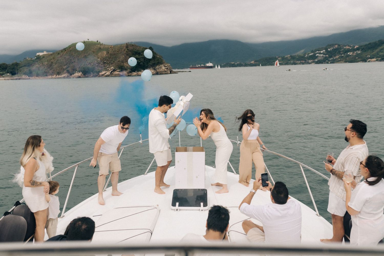 Friends celebrate on a yacht deck with blue smoke and party decorations against a scenic mountain backdrop.