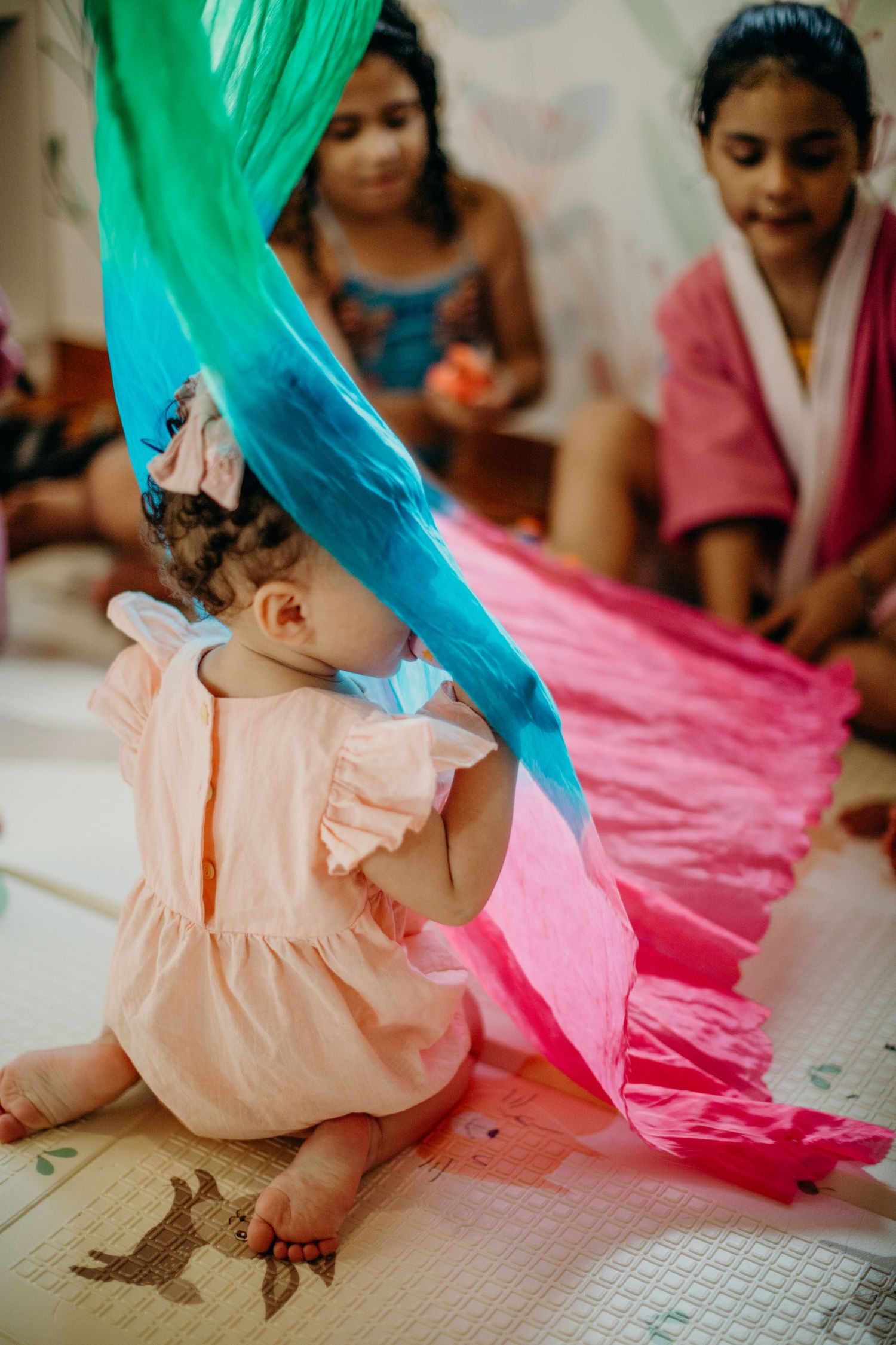 Child in peachy dress sits under colorful fabric drapes on a play mat with animal prints.