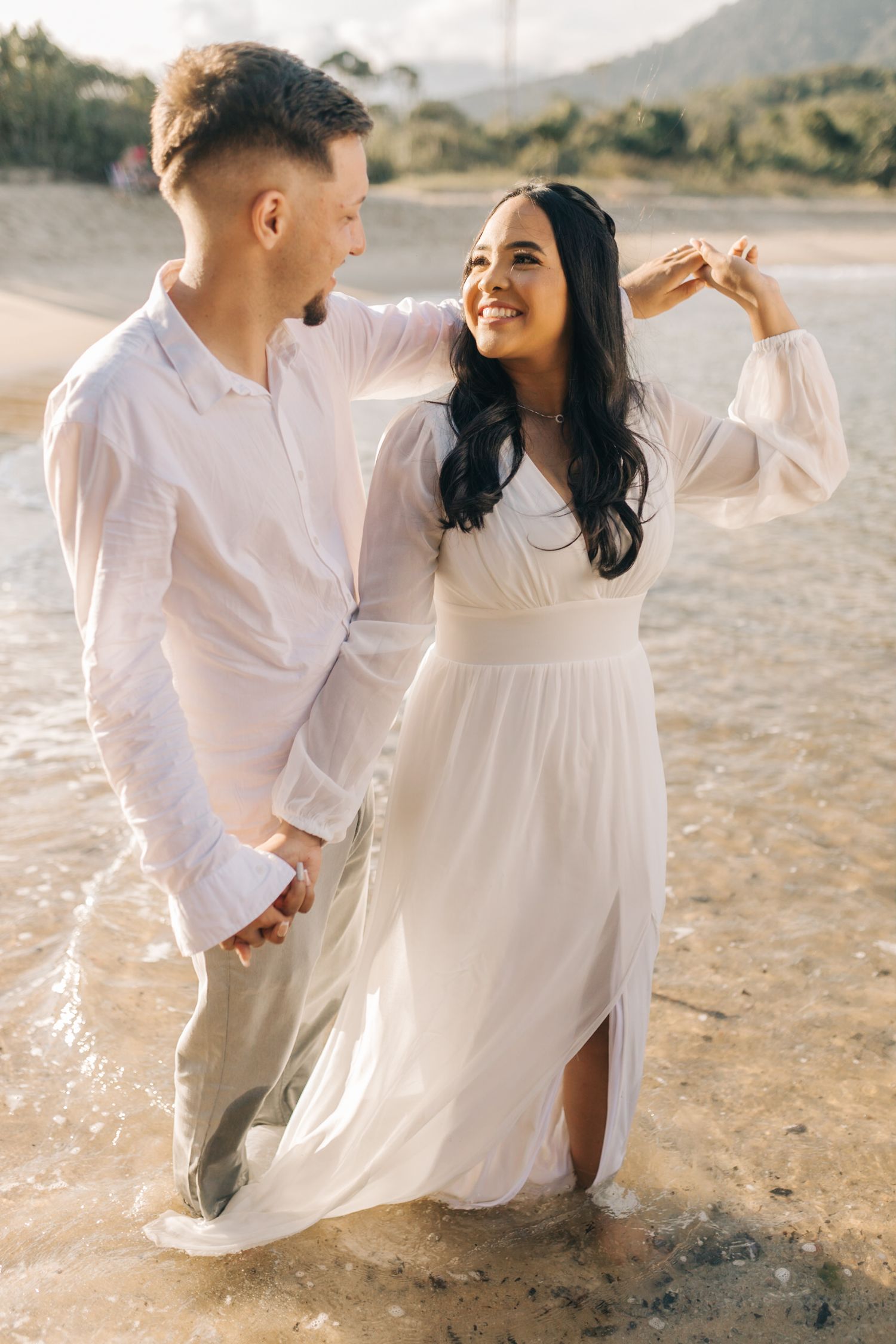 A couple dances playfully on the beach at sunset wearing matching light-colored outfits.