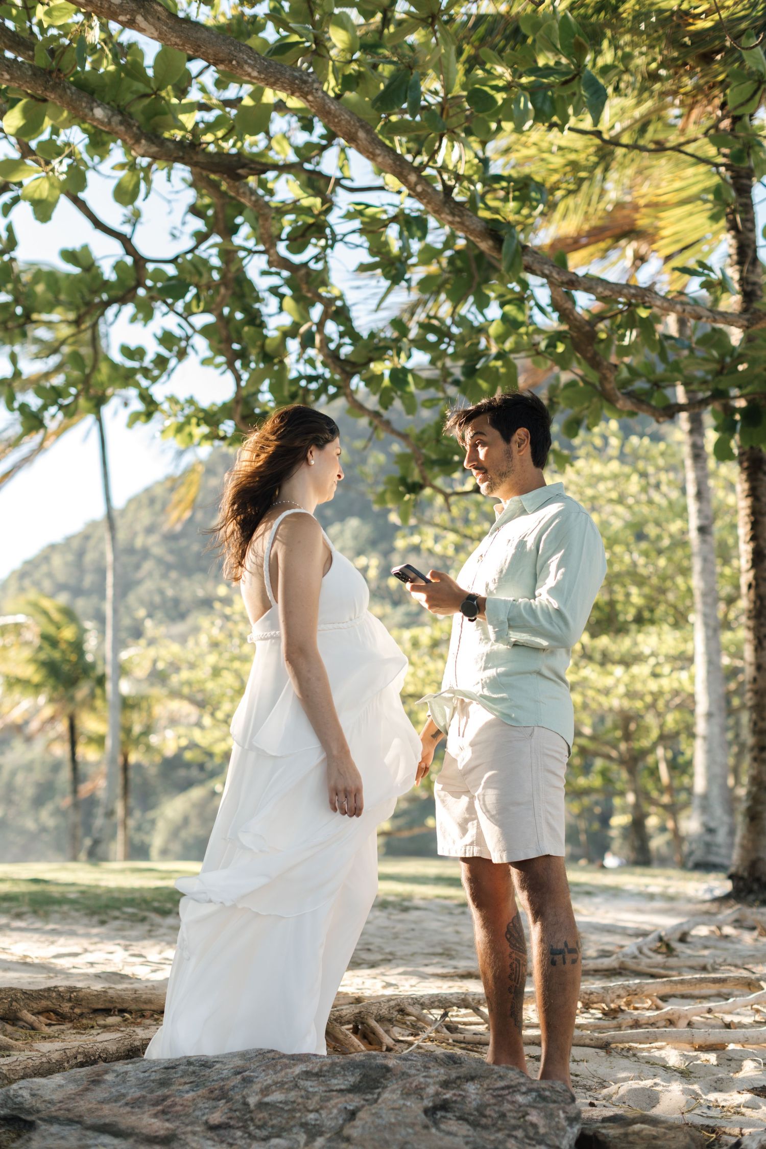 A couple shares a tender moment under green foliage at a tropical beach location.