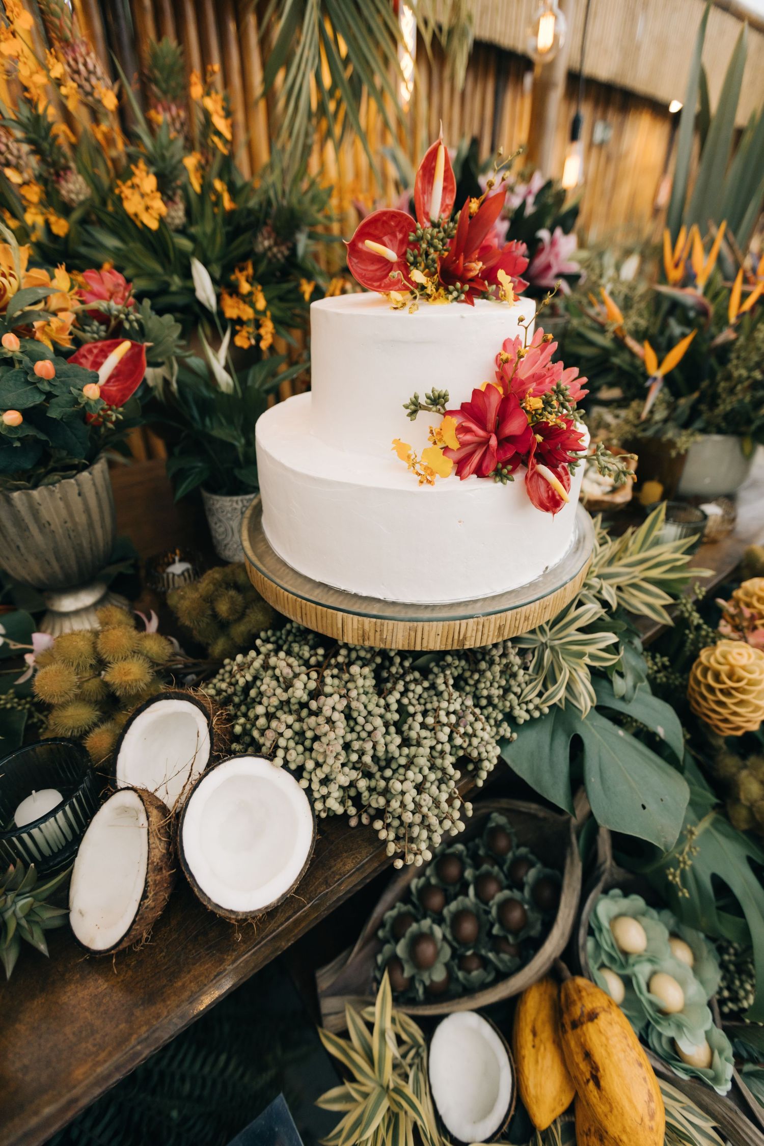 White tiered cake decorated with red tropical flowers on wooden stand with coconuts and greenery.