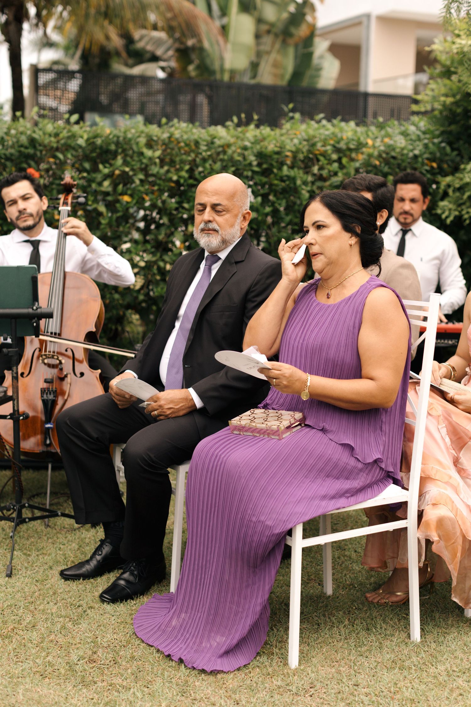 Wedding guests in formal attire seated in white chairs on a lawn with people wearing purple dresses.