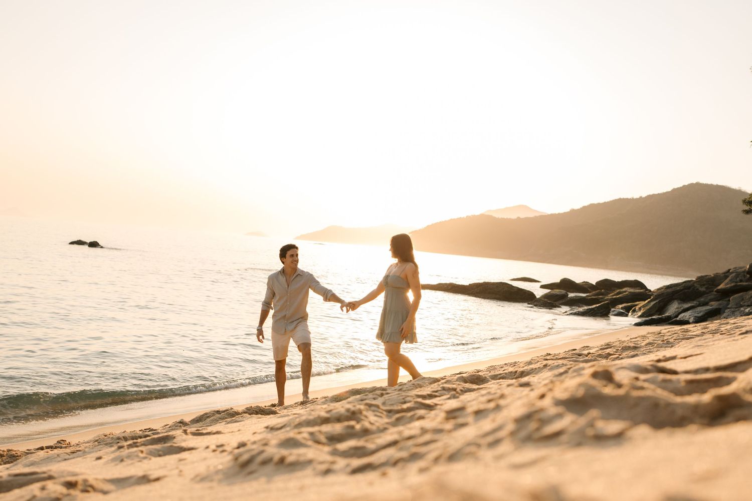 Silhouettes of people walking along a sandy beach during sunset with mountains in the background.