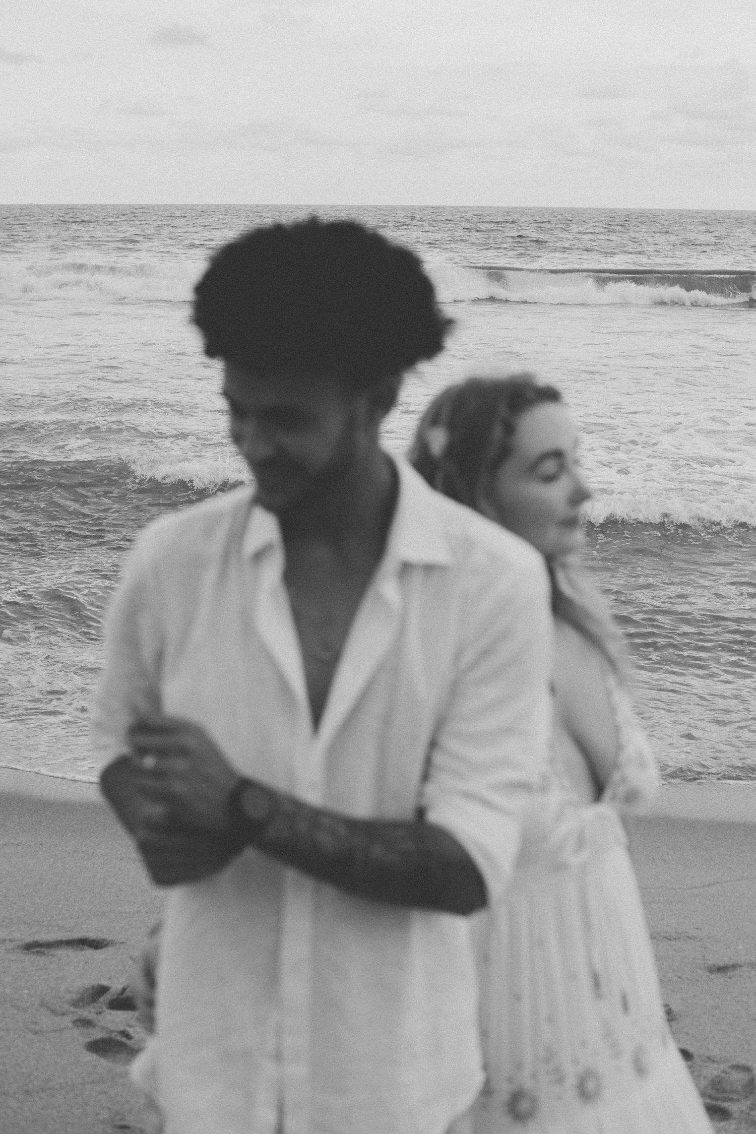 A couple in white clothing embraces playfully on a beach during a black and white photoshoot.