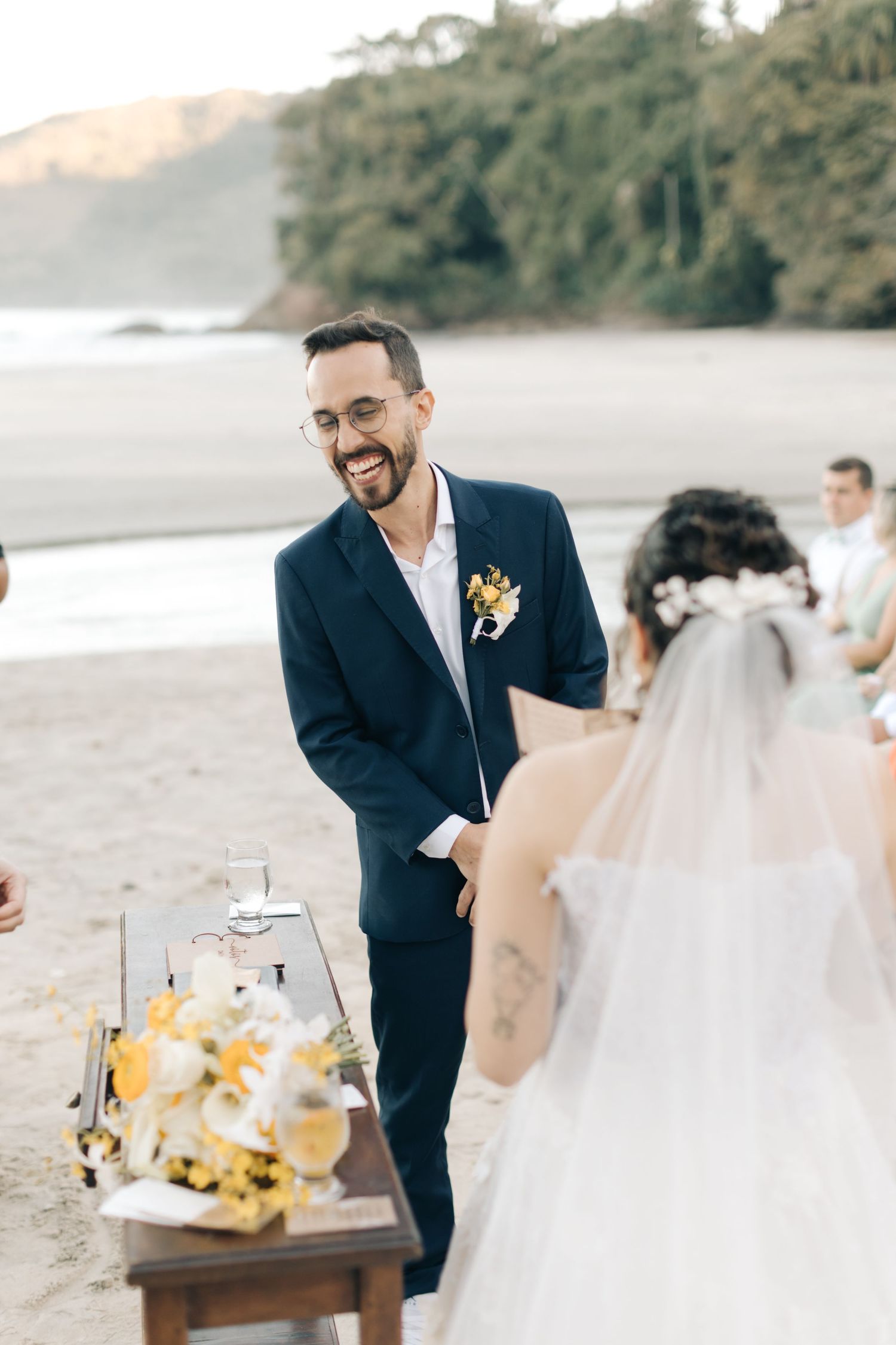 Wedding ceremony on beach with officiant in dark suit and decorative table with yellow flowers.