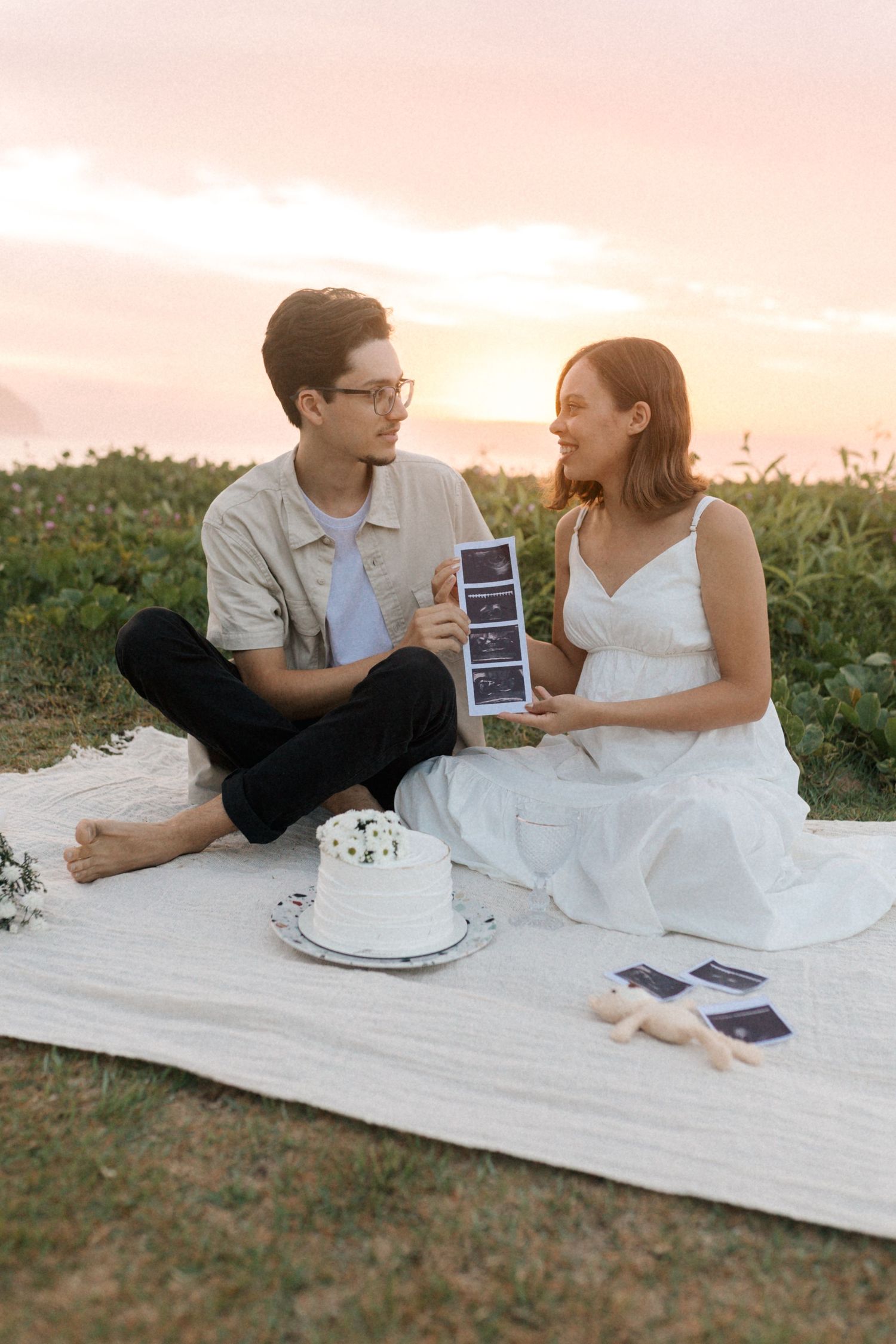 Couple sitting on blanket in field sharing ultrasound photos and celebrating with a white cake at sunset.