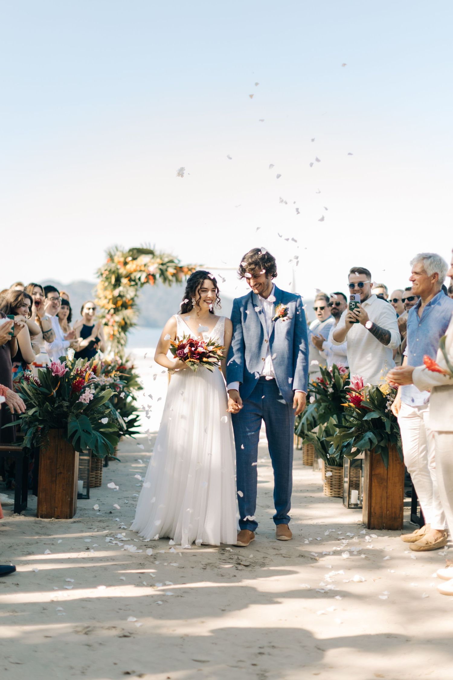 Newlyweds walk down flower-lined beach aisle as guests throw confetti.